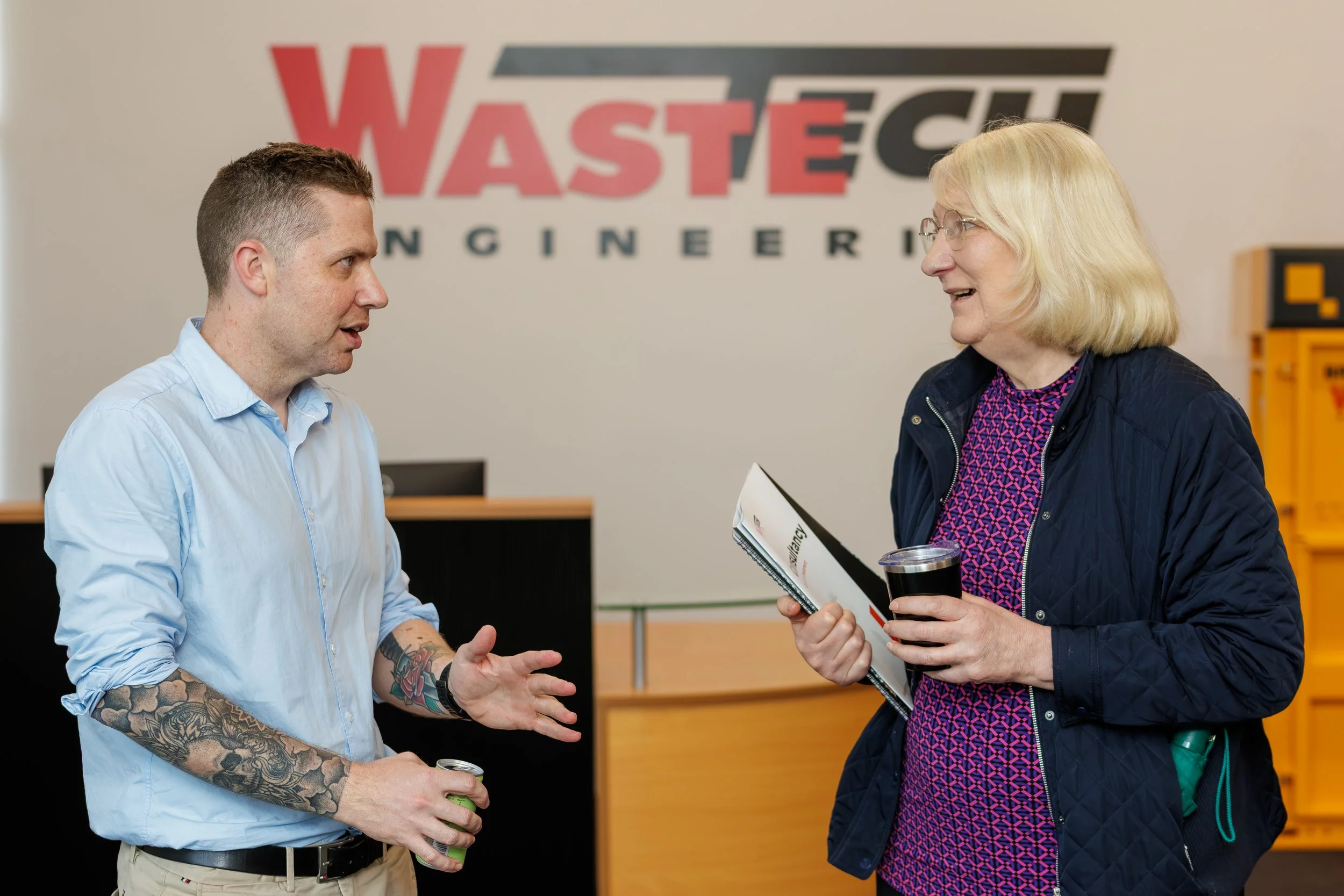 Two people, a man and a woman, are having a conversation at an indoor event. The man has tattoos on his arm and is holding a drink, while the woman is holding a magazine and a drink, smiling. Behind them is a wall with the logo 'WASTE' and other text