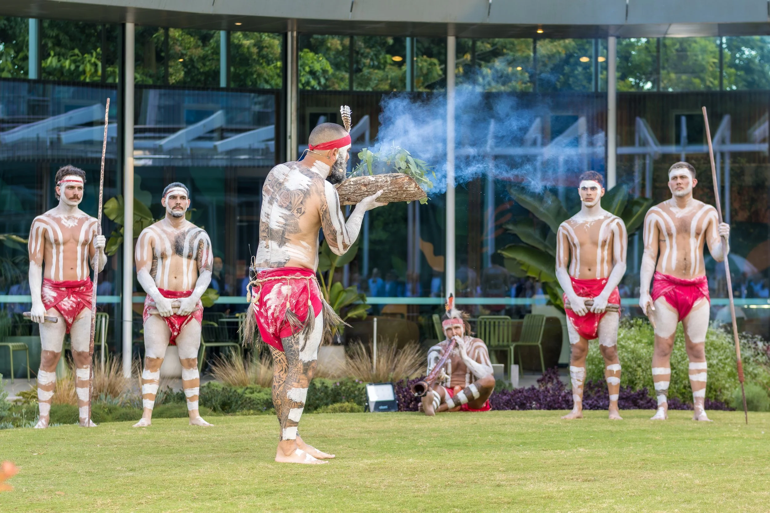 A group of Indigenous men dressed in traditional attire with body paint and red loincloths, participating in a cultural ceremony outdoors with a man holding a wooden tray with smoke, in front of a glass building with trees in the background.