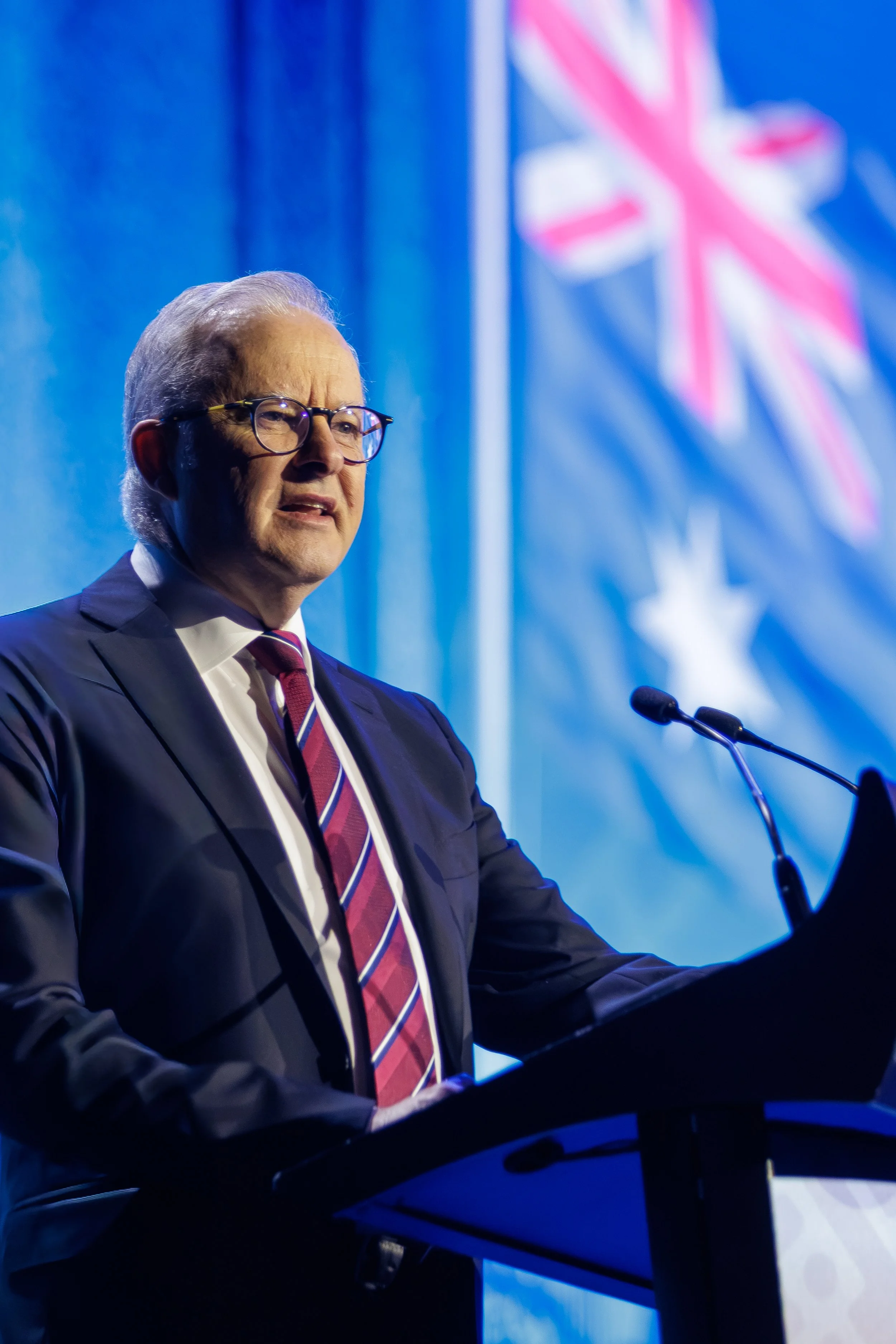 An older man with glasses, wearing a suit and striped tie, speaking at a podium with a microphone in front of a blue background with a pink and white Union Jack flag design.