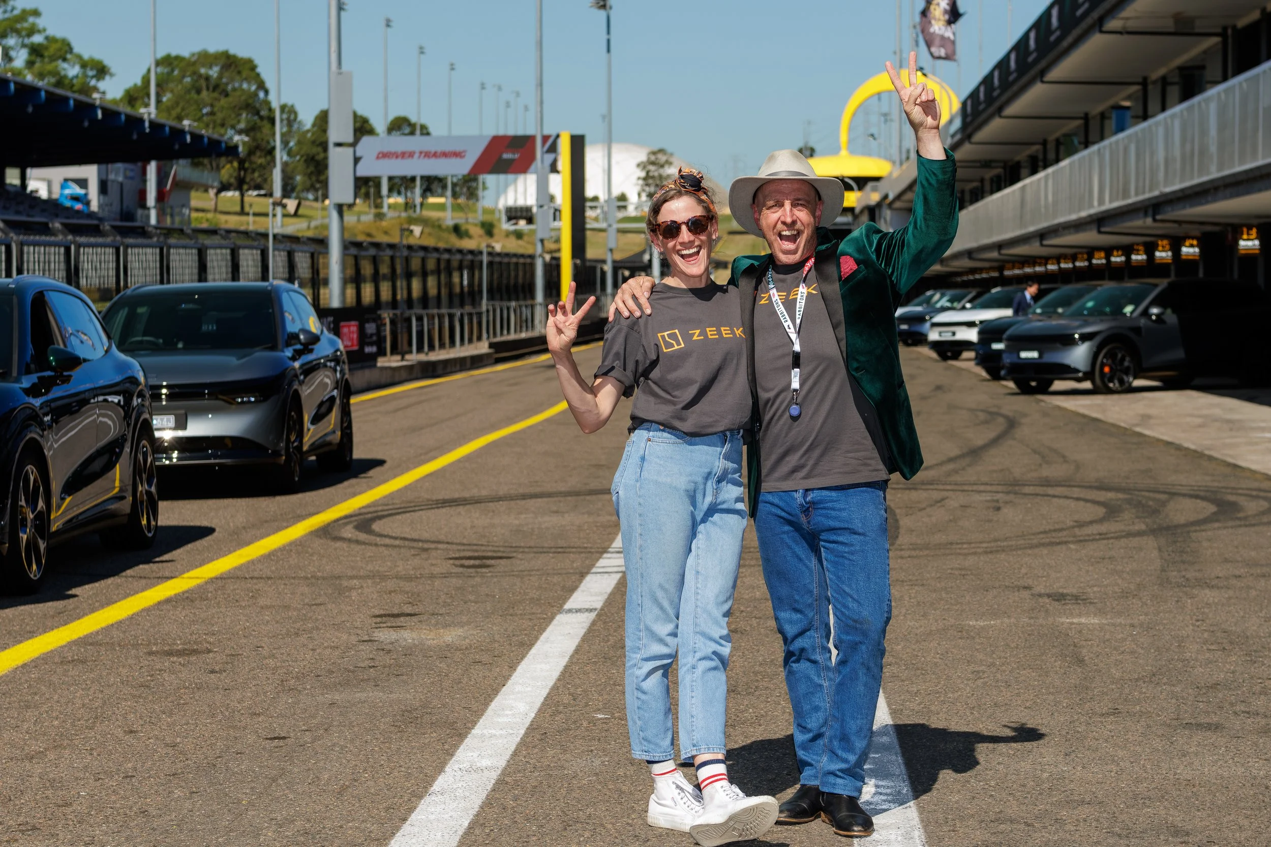 Two people, a woman and a man, smiling and making victory signs at a race track parking lot. The woman is wearing sunglasses and a gray Zeek T-shirt, and the man is wearing a wide-brimmed hat and a green jacket. There are cars parked along the sides 