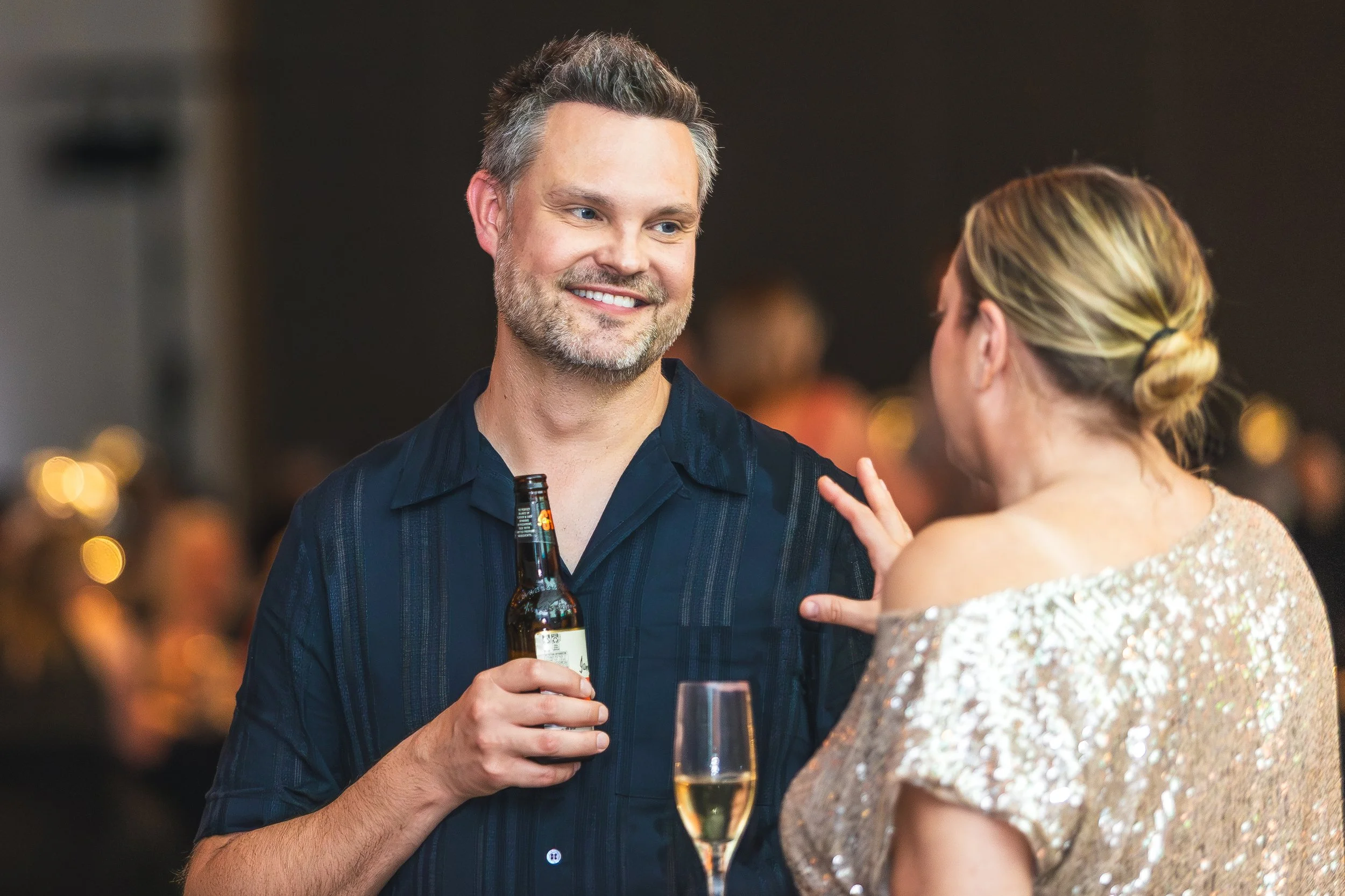 A man with gray hair and a beard smiling while talking to a woman with blond hair in a bun at a social event. The man is holding a beer bottle, and the woman is wearing a sequined dress.