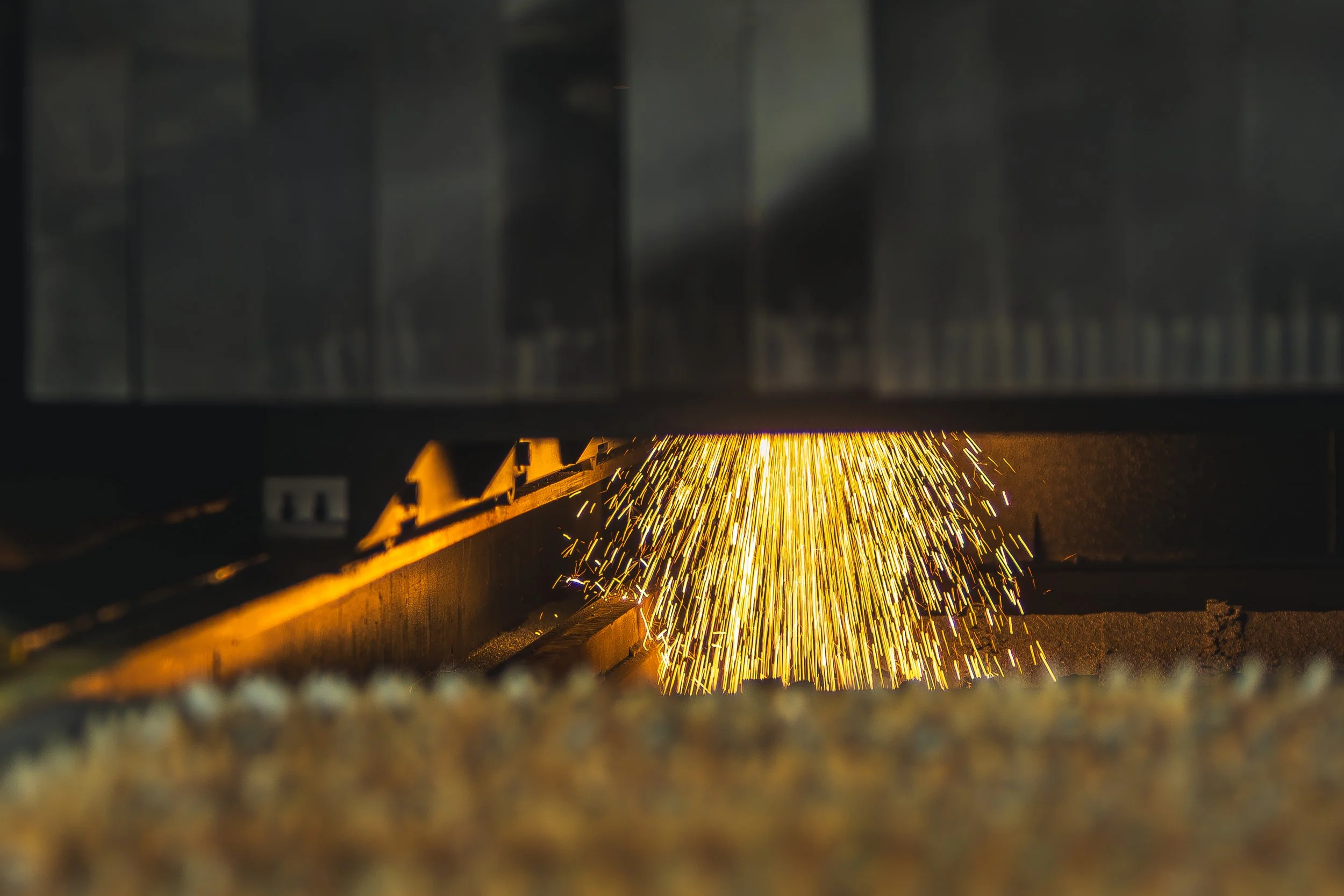 Industrial manufacturing process with sparks flying from metal cutting or welding, seen through a protective barrier.