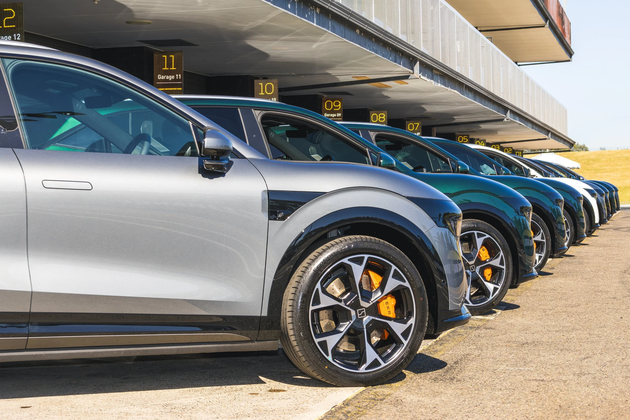 Row of luxury cars parked in a parking lot under a multi-level garage structure with yellow numbered signs indicating garage levels.