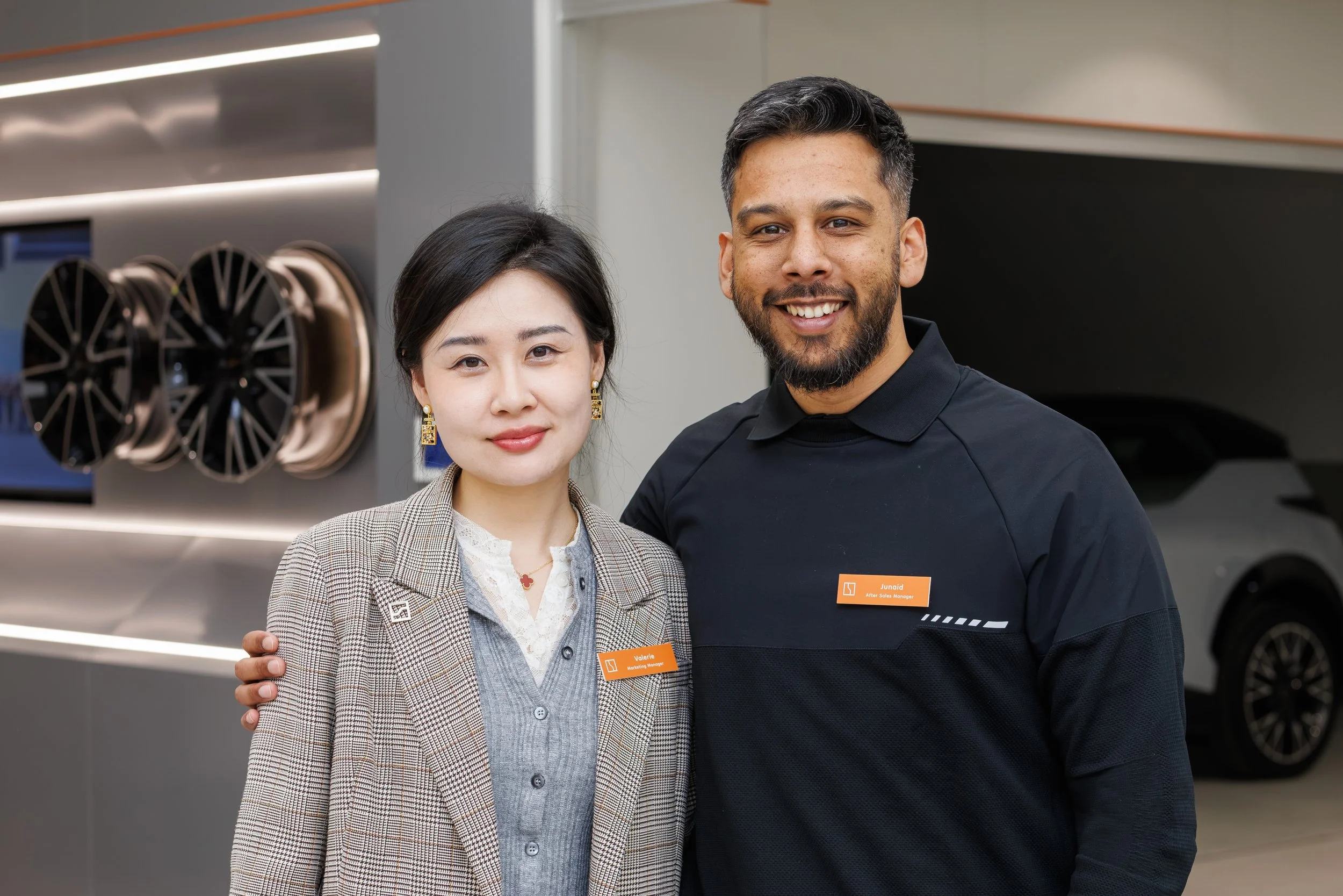 A woman and a man standing close together in a showroom, smiling at the camera. The woman has dark hair, jewelry, and a checked blazer. The man has a beard, short dark hair, a uniform, and an orange name tag reading 'Junaid'.