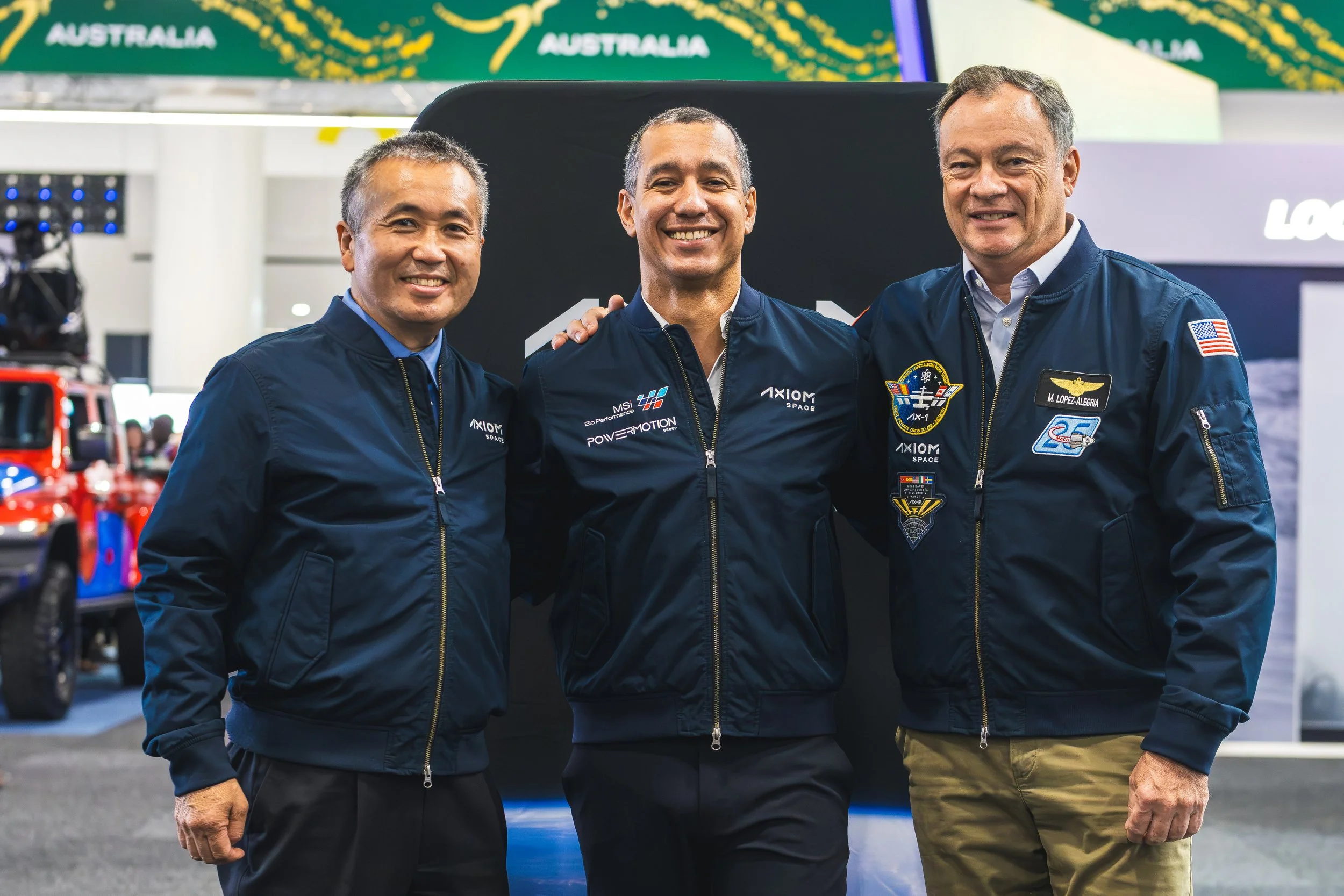 Three smiling men in navy blue jackets standing close together at an indoor event, background showing a display with the word 'Australia' and related graphics.