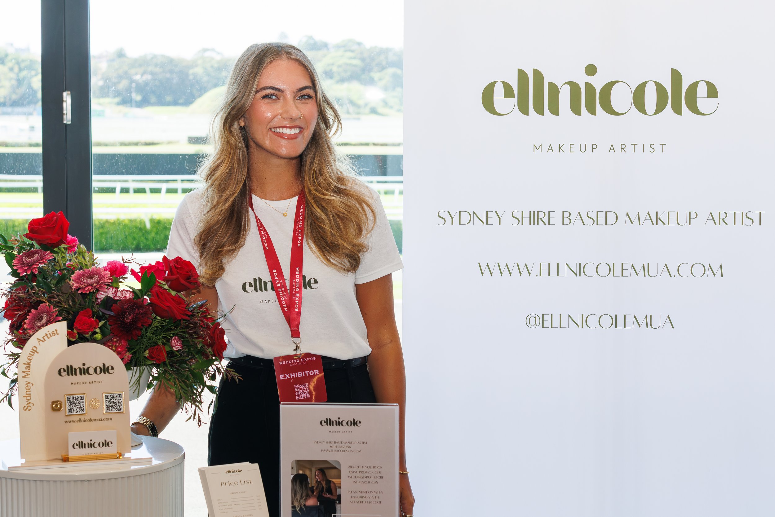 A young woman with long blonde hair smiling at the camera stands next to a table displaying products and flowers at a makeup artist booth. Behind her is a large window with a view of outdoors and a sign promoting Ella Nicole, a makeup artist based in