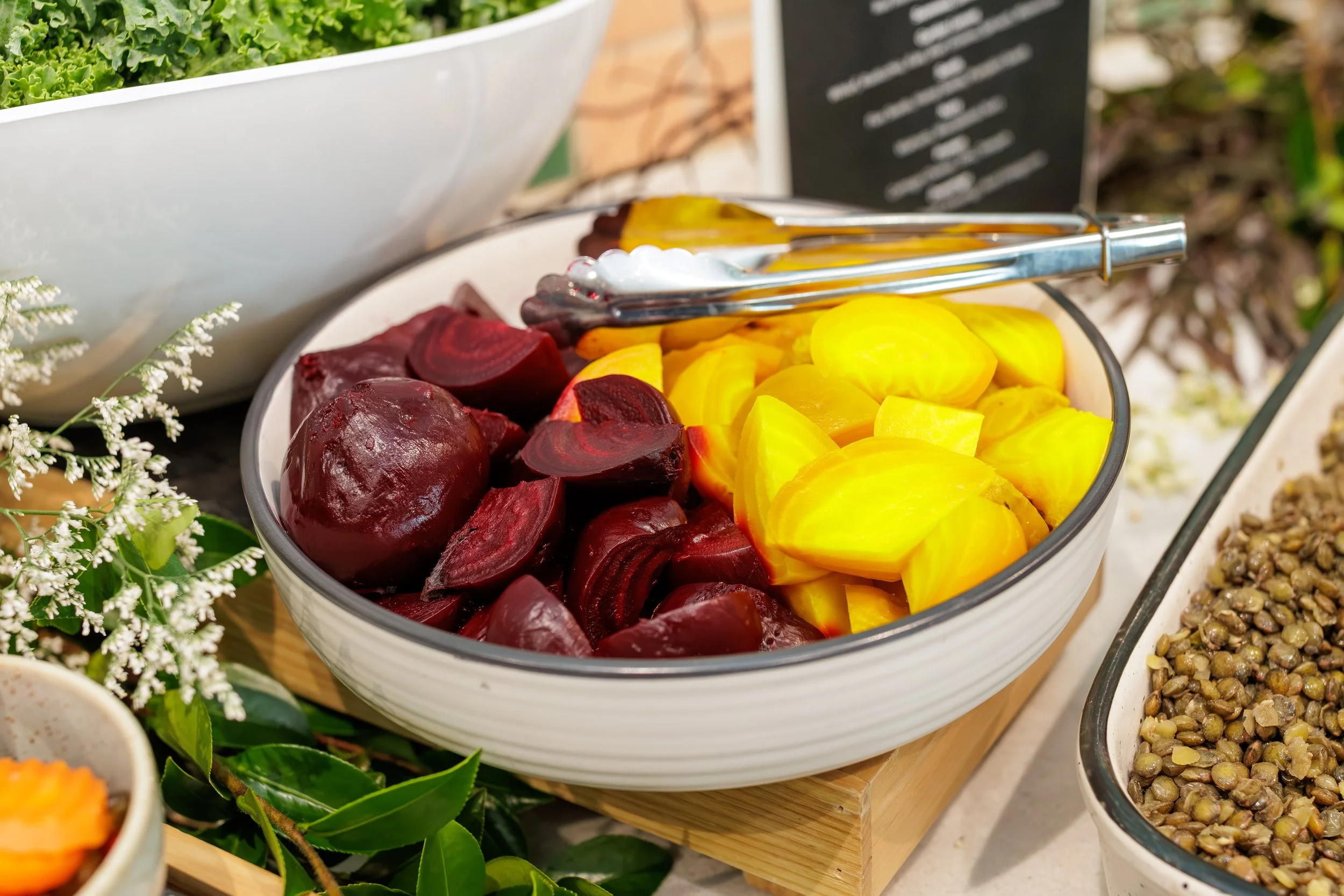 A bowl of sliced beets and sliced yellow peaches, with tongs on top, at a salad bar or buffet.