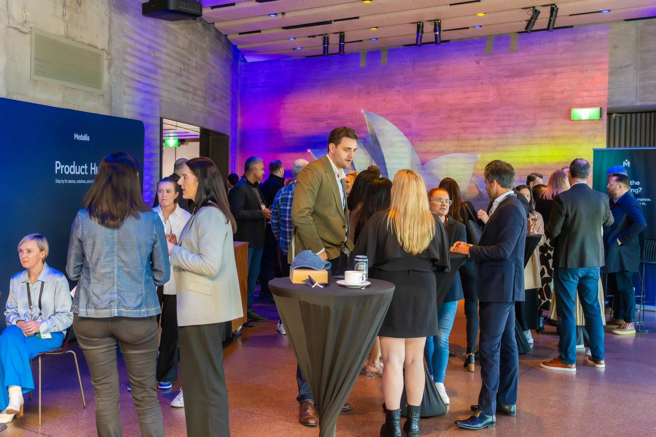 A group of people attending a conference or networking event in a large room with a high ceiling. There are several standing tables with drinks, and a screen on the left displays 'Product Hi' with smaller text underneath. The room is decorated with c