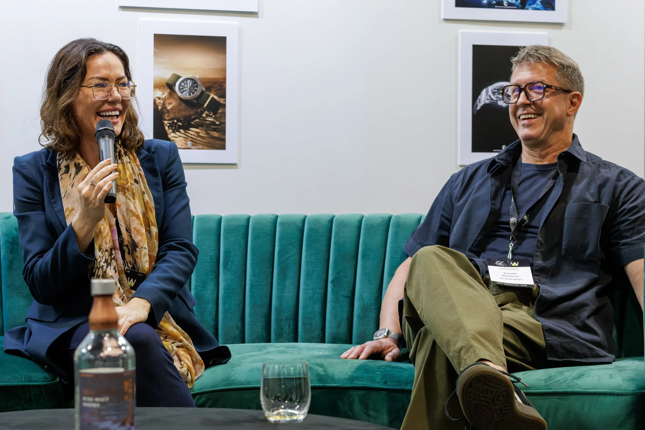 Two people, a woman and a man, are sitting on a green velvet couch at an indoor event. The woman has shoulder-length brown hair, glasses, and is holding a microphone while smiling. She is wearing a blue blazer and a patterned scarf. The man has short