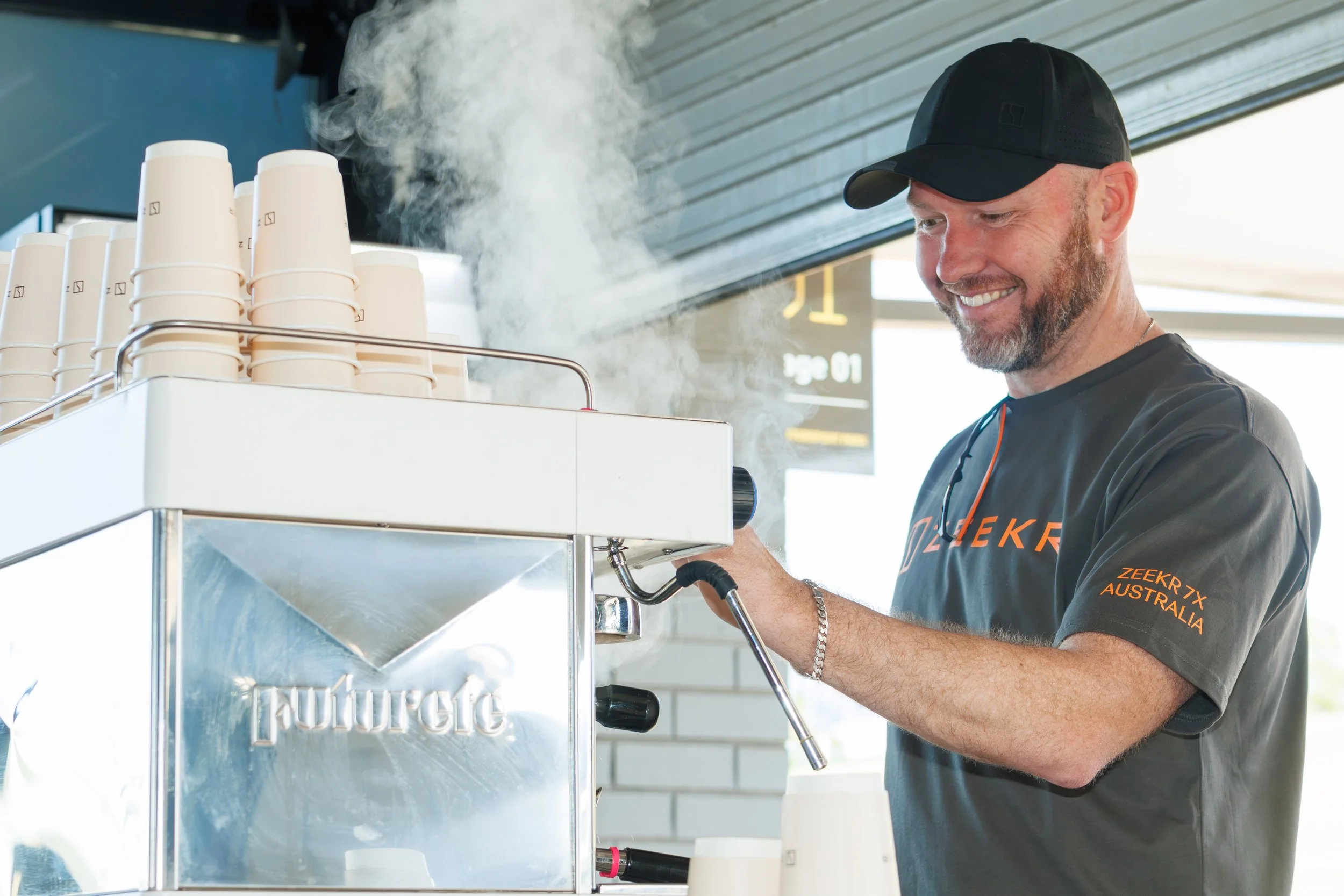 A man smiling while making coffee at a coffee machine in an indoor setting.