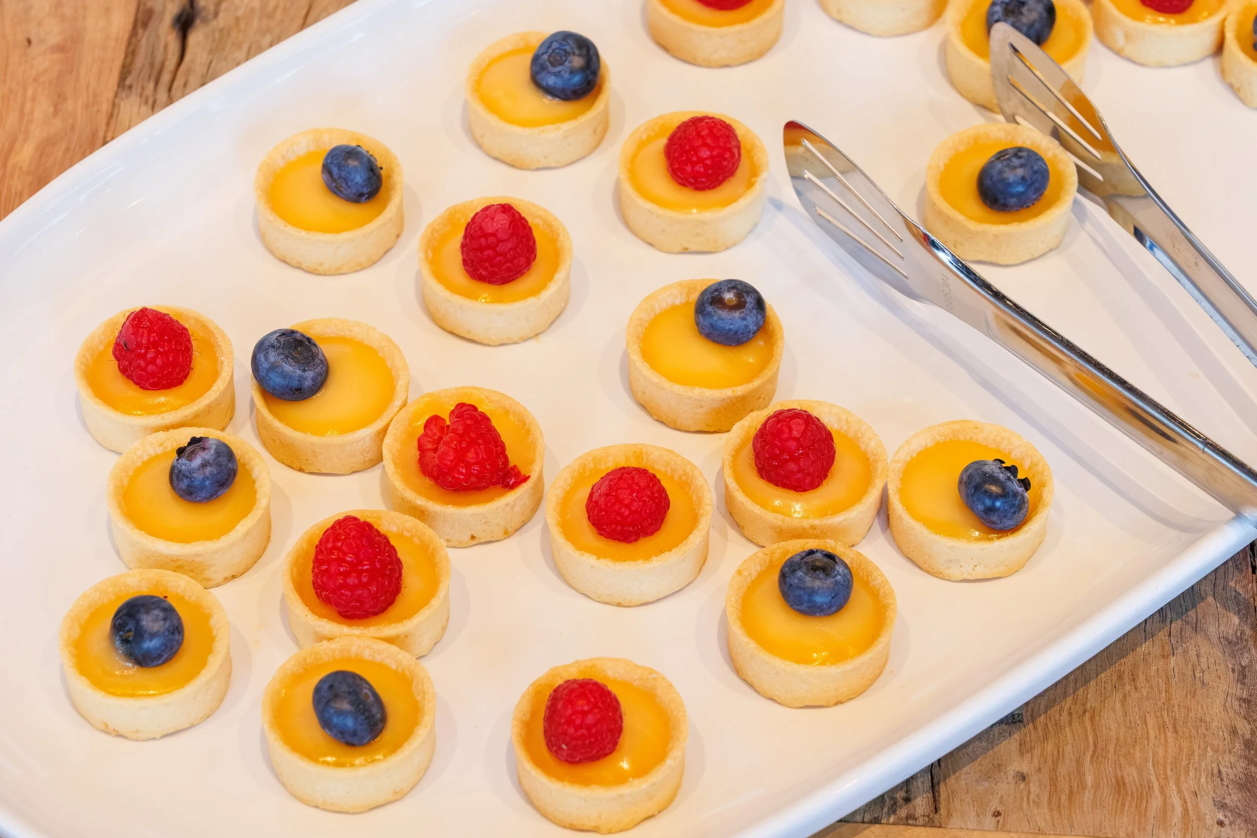 Small fruit tartlets on a white serving platter, each topped with blueberries or raspberries.