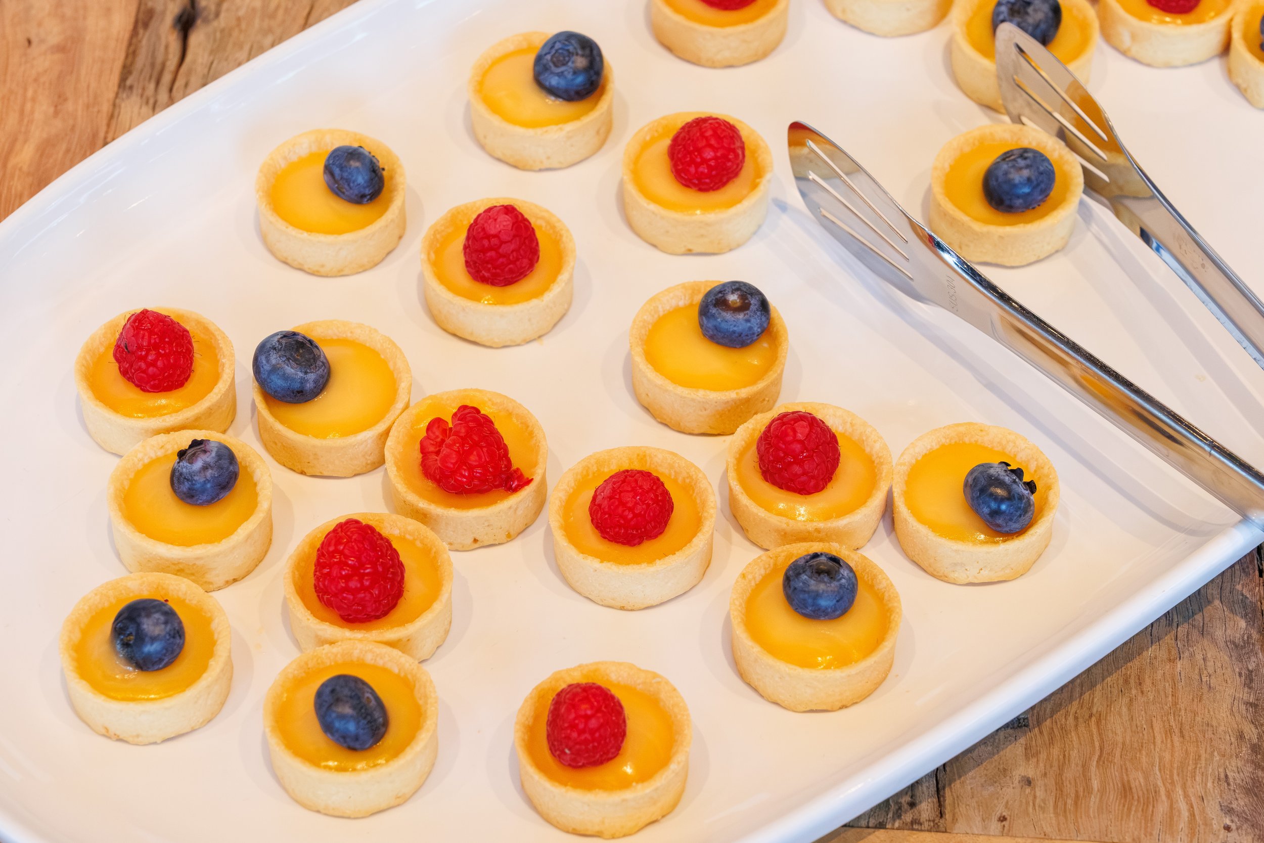Small fruit tartlets on a white serving platter, each topped with blueberries or raspberries.