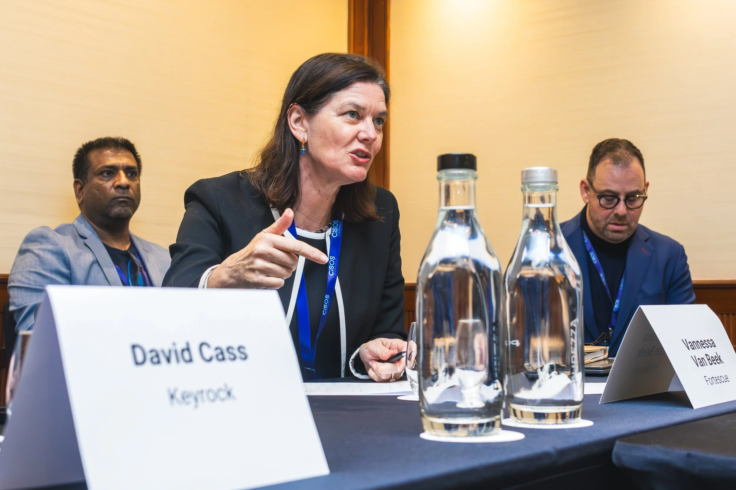 A woman speaking at a panel discussion with two men beside her. The woman has dark hair and is wearing a black blazer, with a CISOS conference lanyard around her neck. The panel table has bottled water and nameplates for each speaker, with the names 