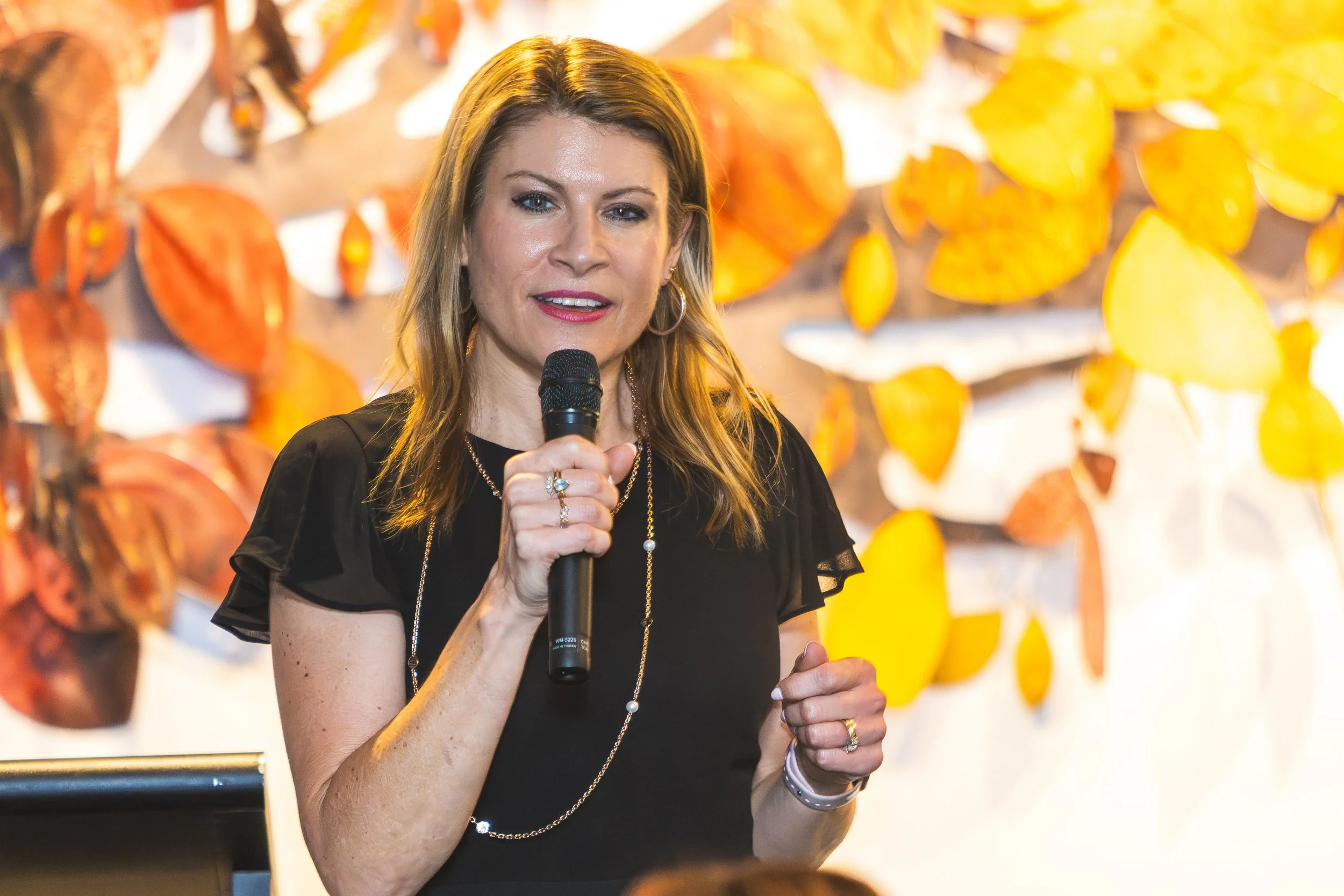 A woman with long blonde hair holding a microphone speaking at an event, with an art display of orange, yellow, and red hanging objects in the background.