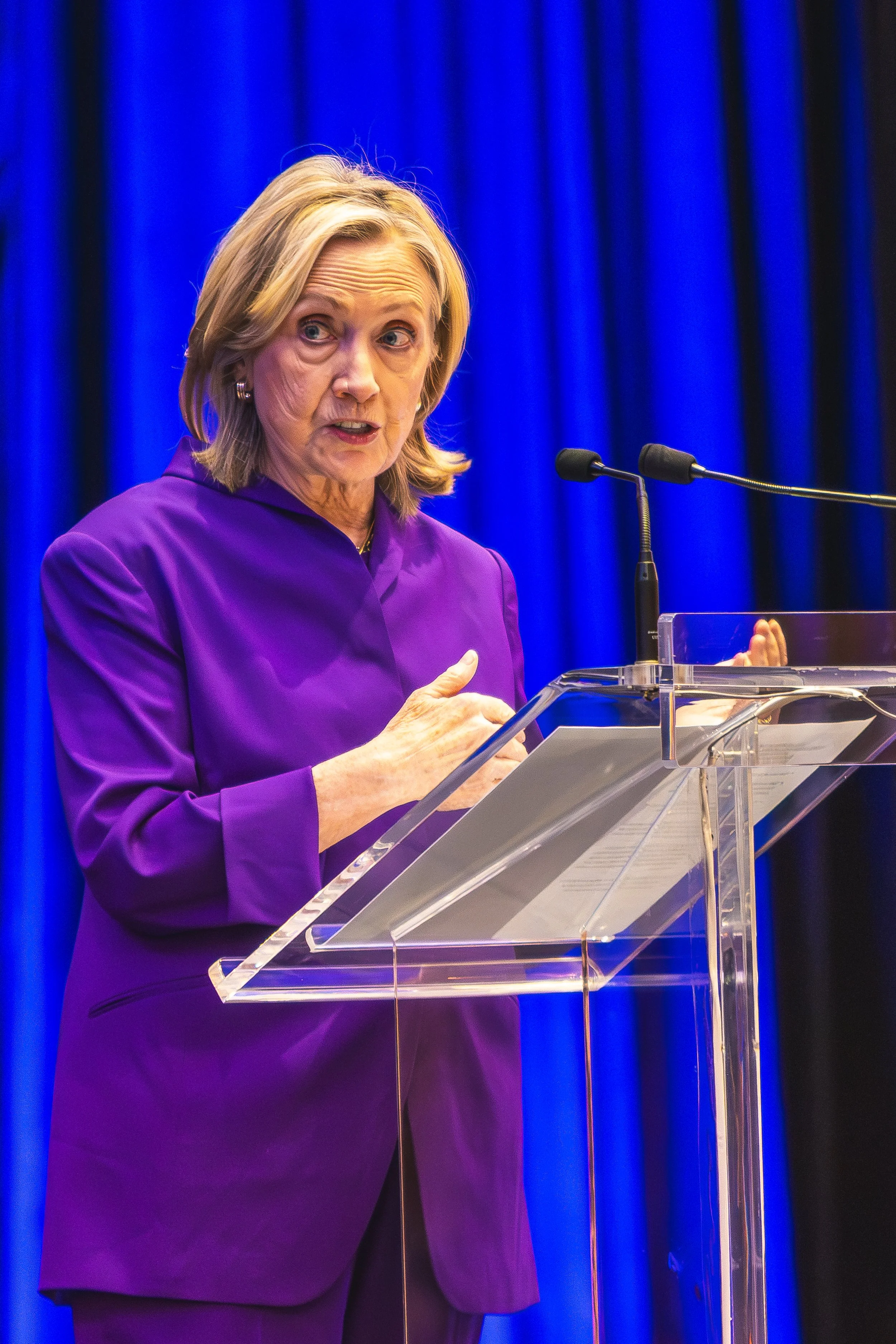A woman with blonde hair wearing a purple blazer, speaking at a podium with a microphone, against a blue curtain background.