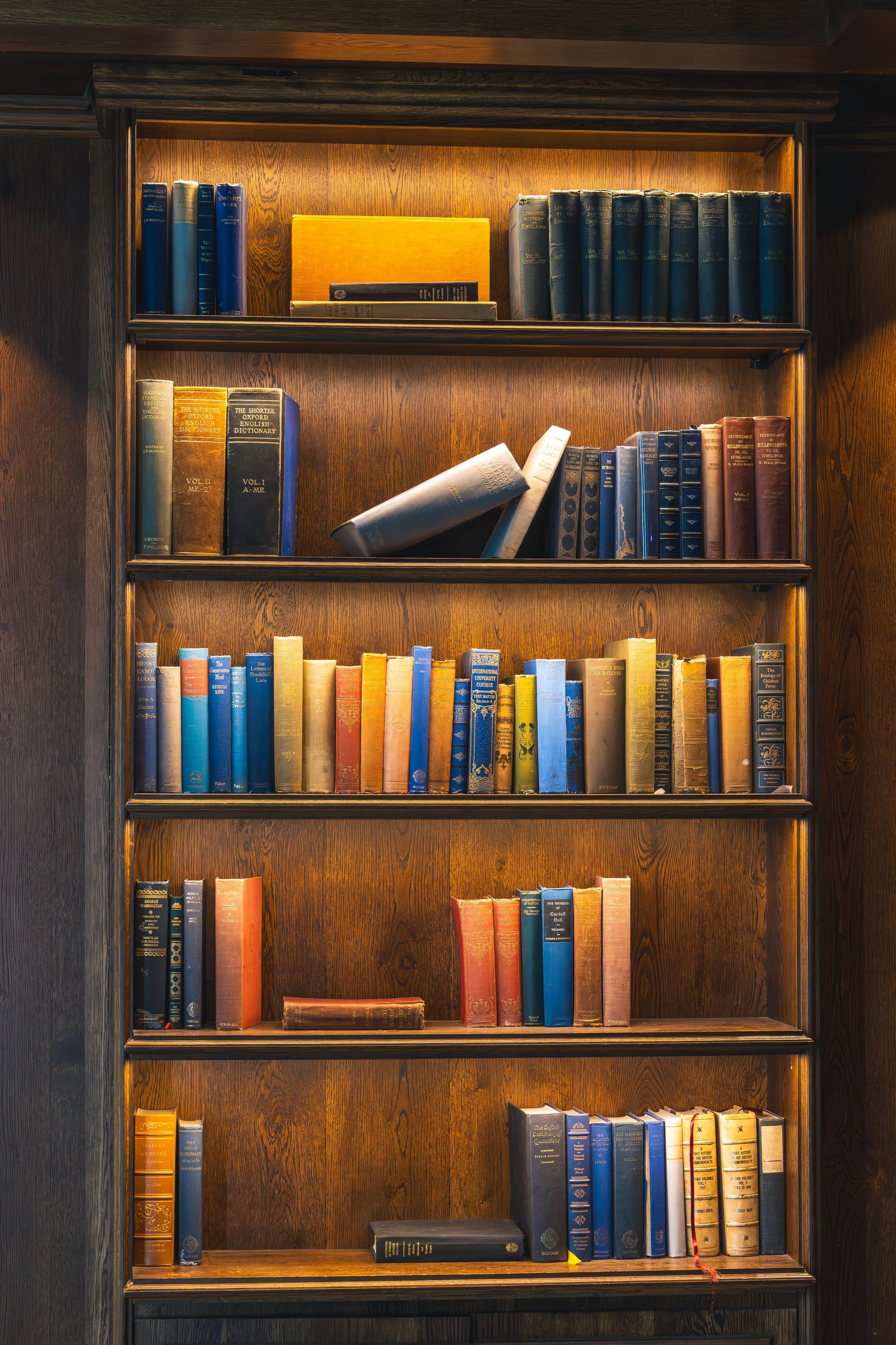 A wooden bookshelf filled with various hardcover books organized in multiple shelves, some leaning, on a dark wood background.