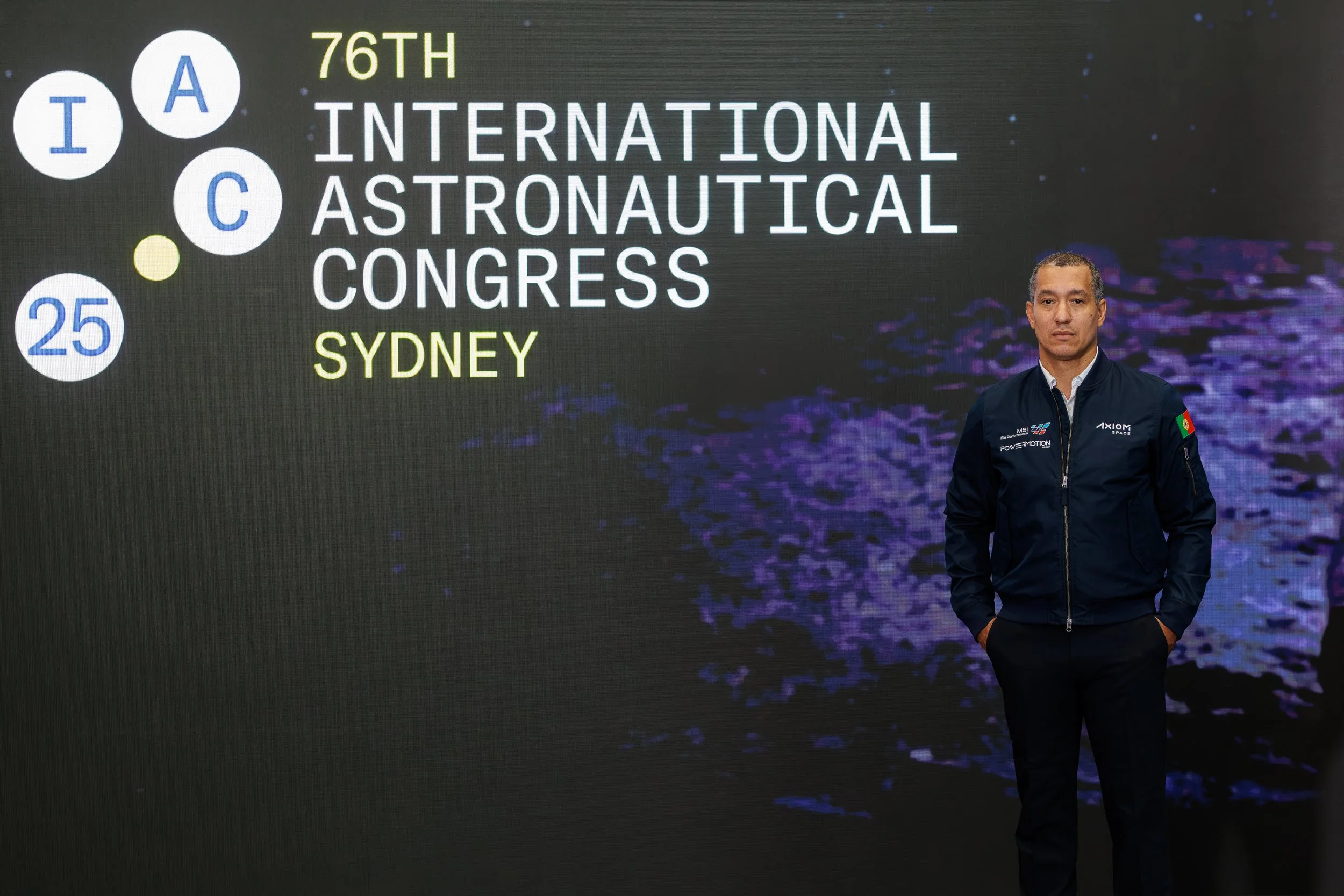 A man standing in front of a large digital display with the text '76th International Astronautical Congress Sydney' and various logo symbols.