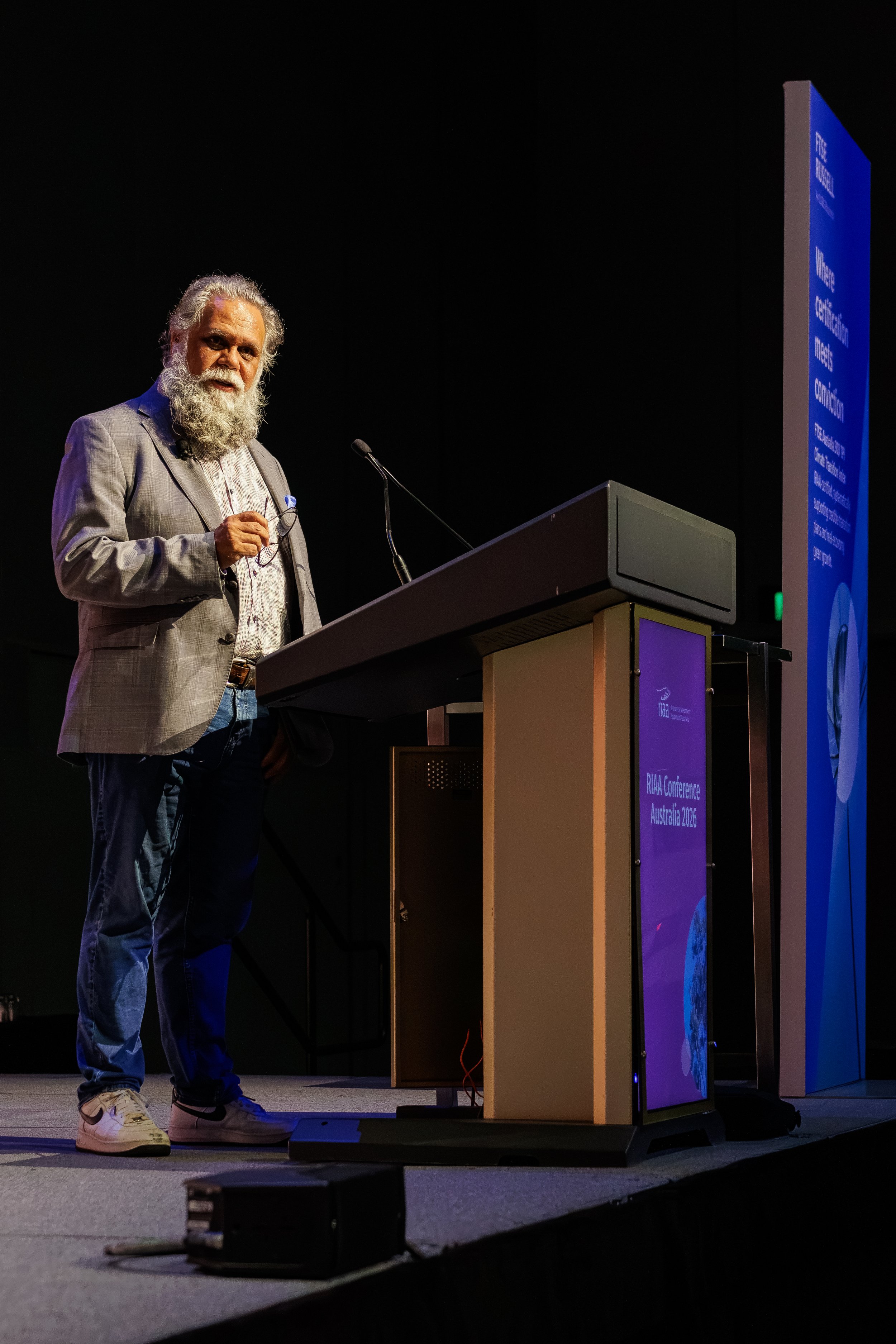 A man with a long gray beard and wearing a gray blazer, beige shirt, and jeans standing on stage at a conference, holding glasses, with a large blue and purple presentation screen behind him that reads 'RIAC Conference Australia 2026'.