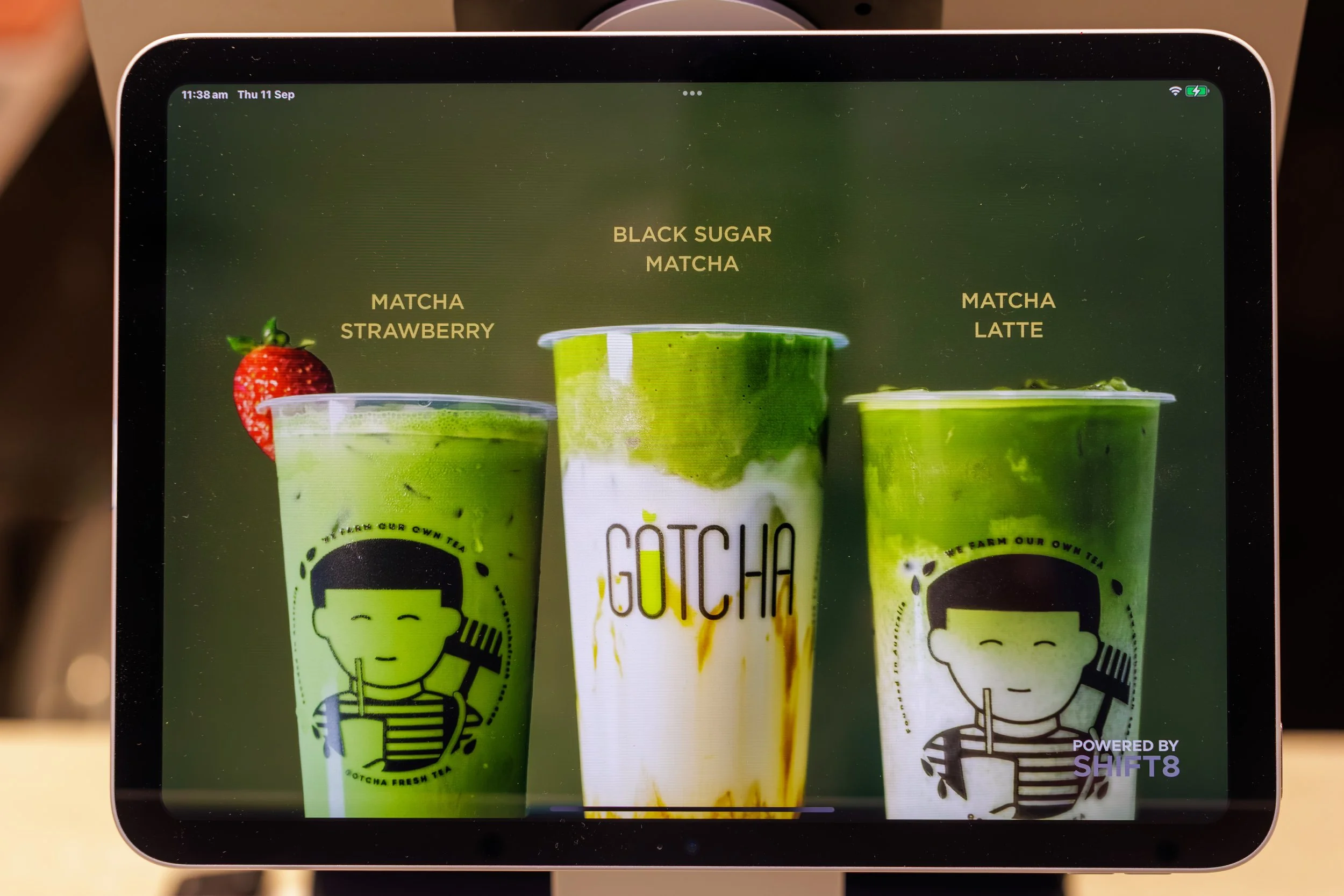 Three cups of matcha beverages with different flavors labeled 'Matcha Strawberry,' 'Black Sugar Matcha,' and 'Matcha Latte,' each in clear plastic cups with green tea drinks inside. A strawberry is on the rim of the left cup.