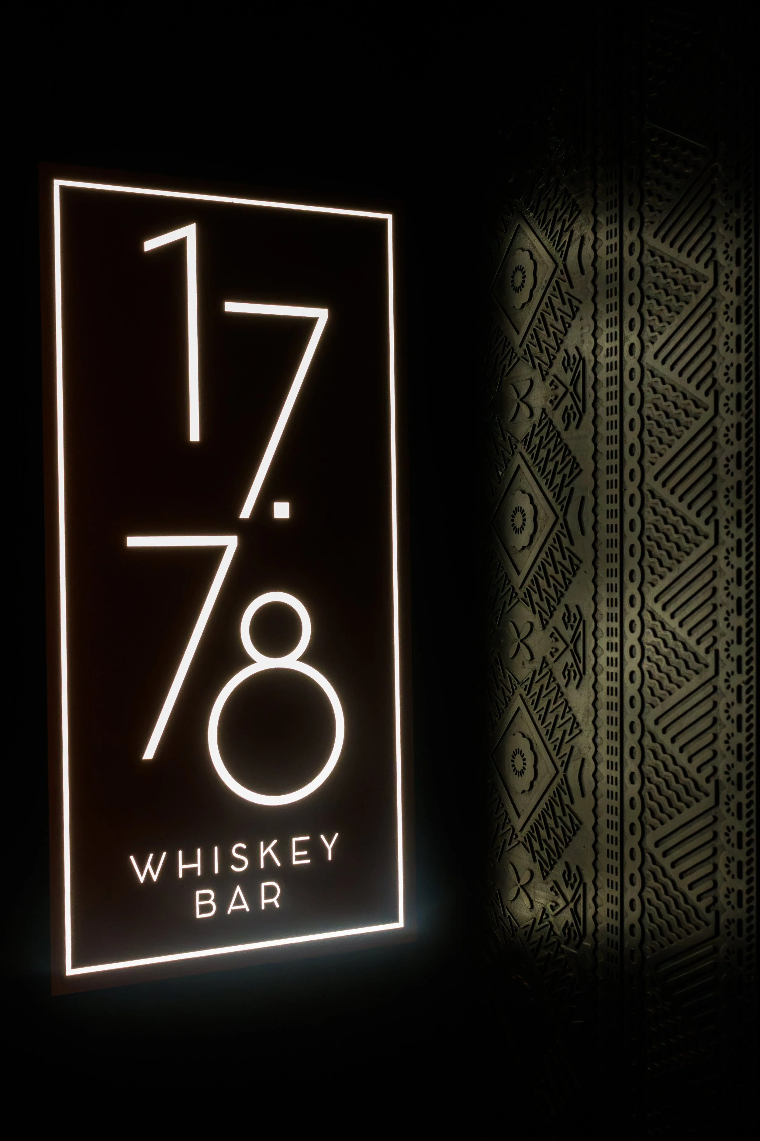 Neon sign for Whiskey Bar with the words, '17', '7', and '8' displayed prominently, and decorative patterned wall on the right side.