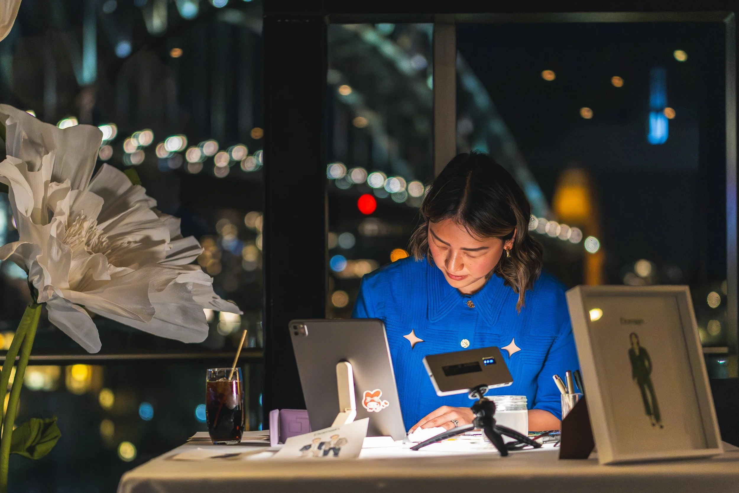 A woman in a blue dress with star-shaped cutouts working at a table in a dimly lit environment with bokeh lights in the background. There is a white flower, a glass of dark beverage, and electronic devices on the table.