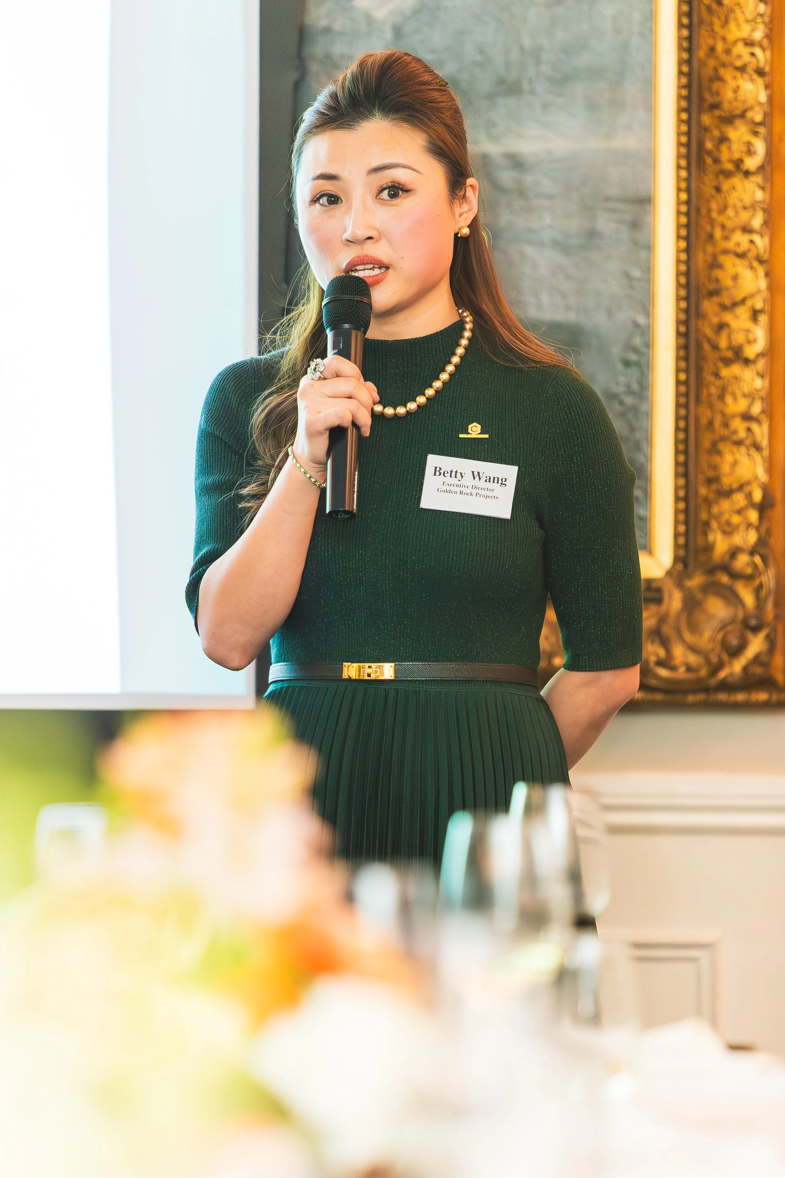 A woman holding a microphone, wearing a black dress and pearl jewelry, standing in a room with ornate decorations, possibly giving a speech or presentation.