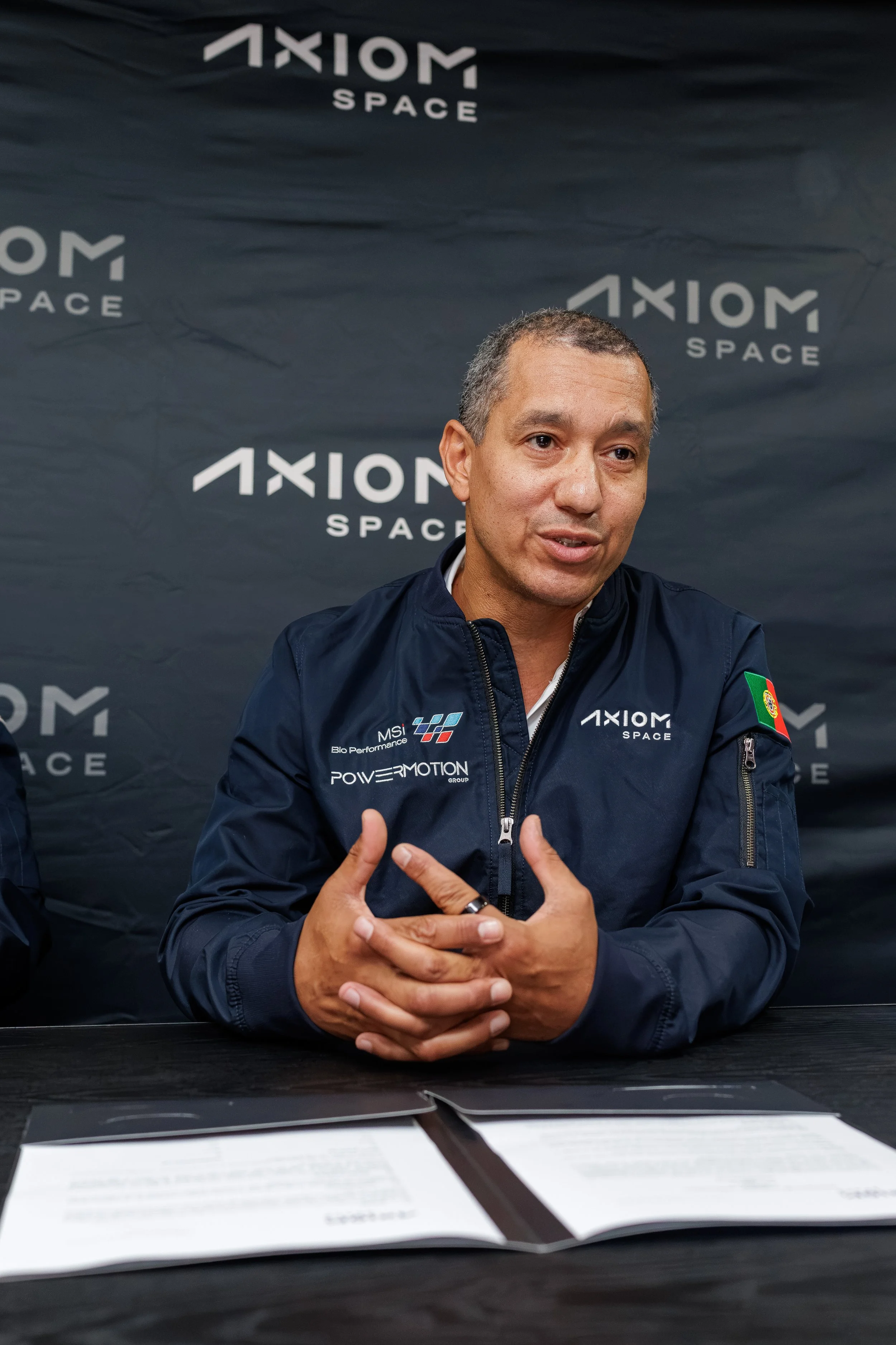 A man speaking at a press conference or interview, seated in front of a black backdrop with 'Axiom Space' logos, wearing a dark jacket with sponsor logos, gesturing with his hands.