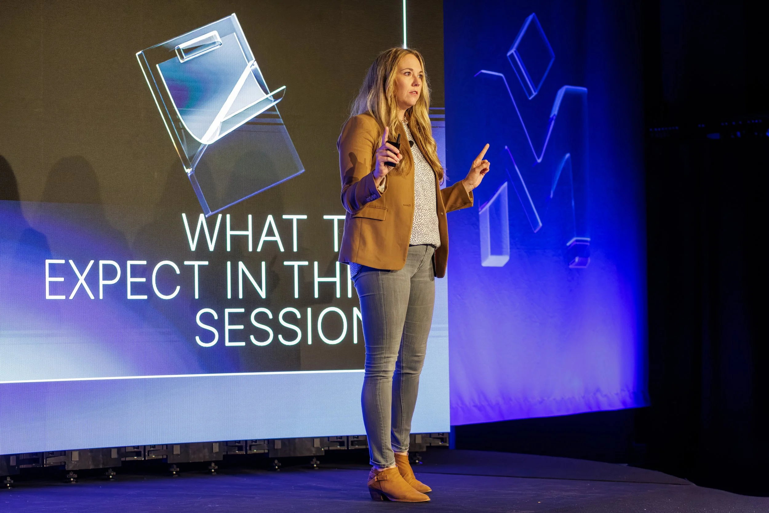 A woman in a brown blazer, beige pants, and tan boots is speaking in front of a large digital display showing the text 'What to Expect in the Session' and a graphic design. She holds a clicker or remote in her right hand and gestures with her left ha