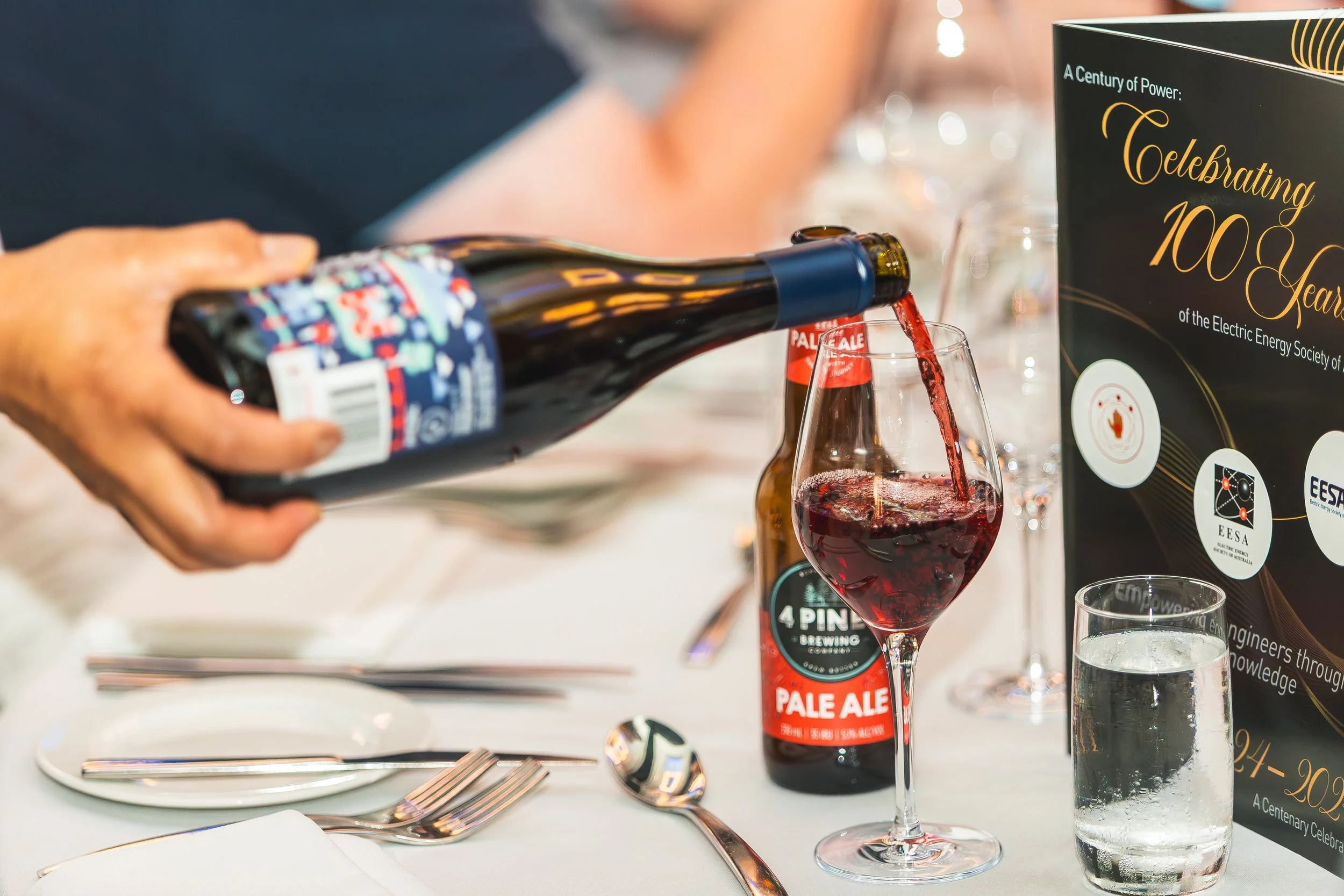 Person pouring red liquid from a bottle of Pale Ale into a wine glass at an event, with a sign celebrating 100 years of energy and tables set with utensils in the background.