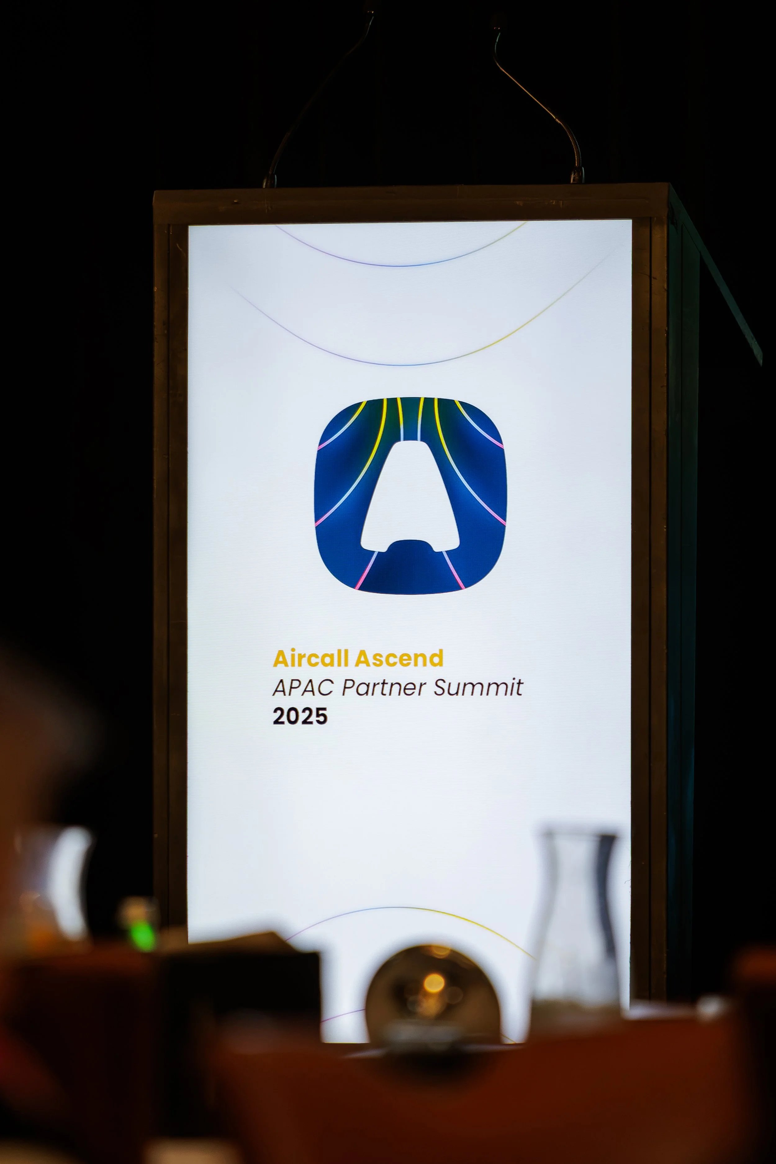 A digital screen displaying information about the Aircall Ascend APAC Partner Summit 2025. The screen shows a logo with a stylized face outline and colorful lines.