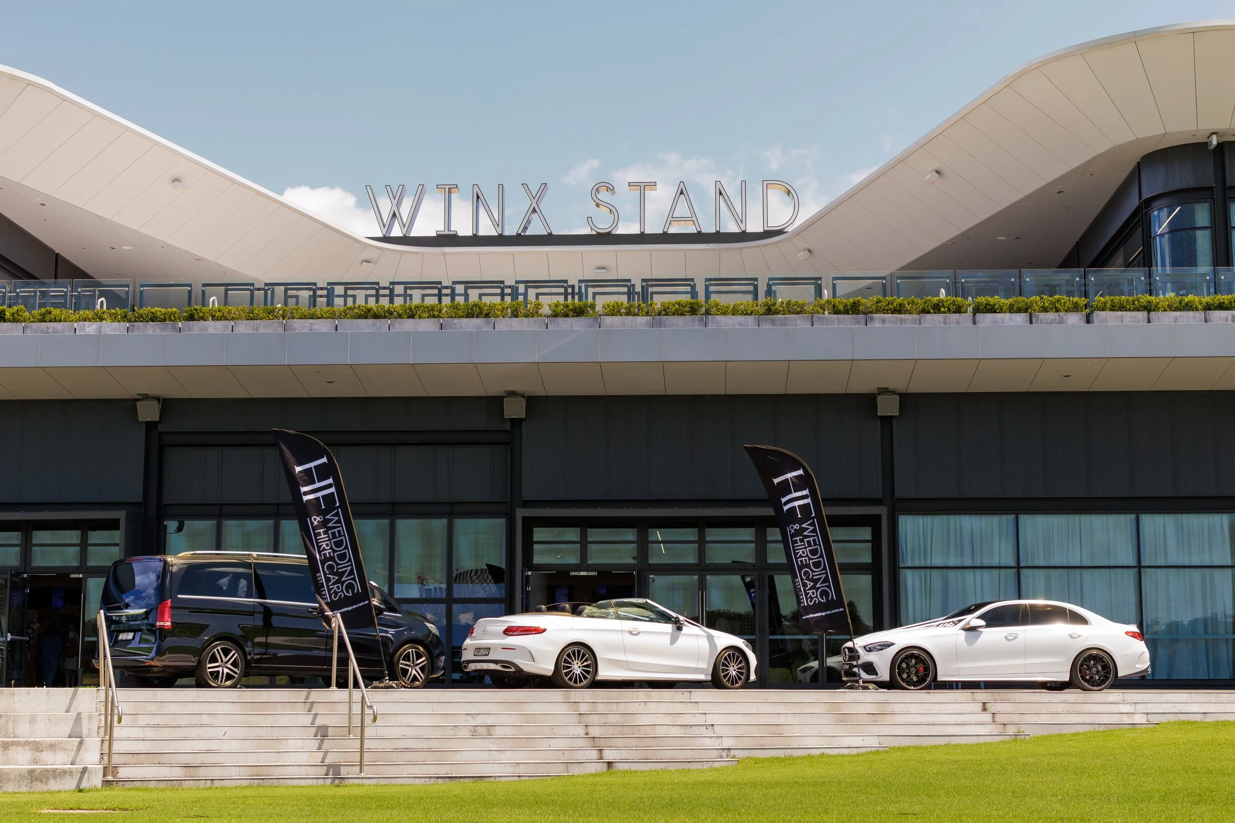 Exterior of Winx Stand building with cars parked in front and wedding, wedding, hire cars flags.