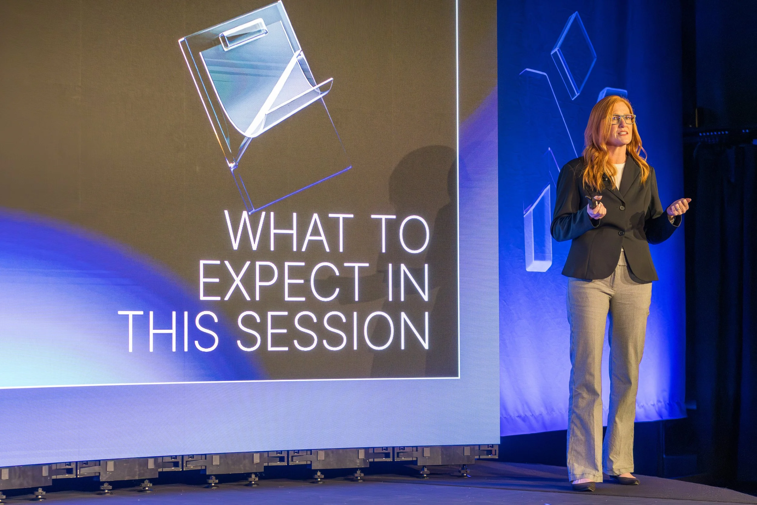 A woman with red hair and glasses giving a presentation on stage, next to a large screen displaying the words 'What to Expect in This Session' and a graphic of a lightbox