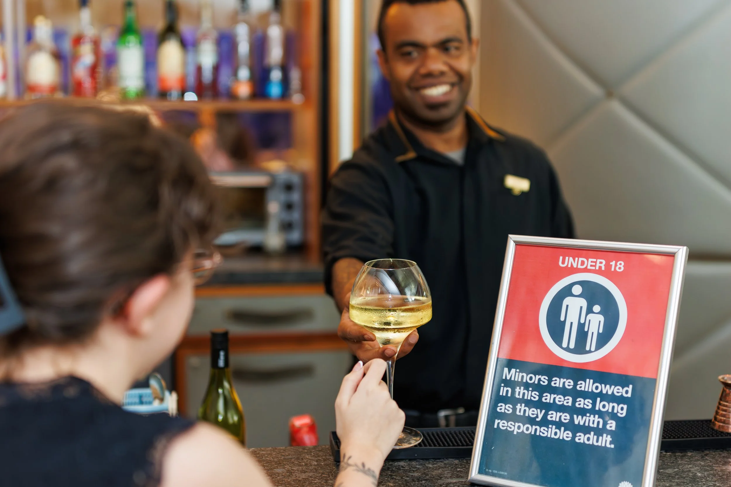 Bartender smiling and holding a glass of white wine while serving a customer at a bar. A sign on the bar reads 'Minors are allowed in this area as long as they are with a responsible adult.' The background shows a shelf with various bottles of alcoho