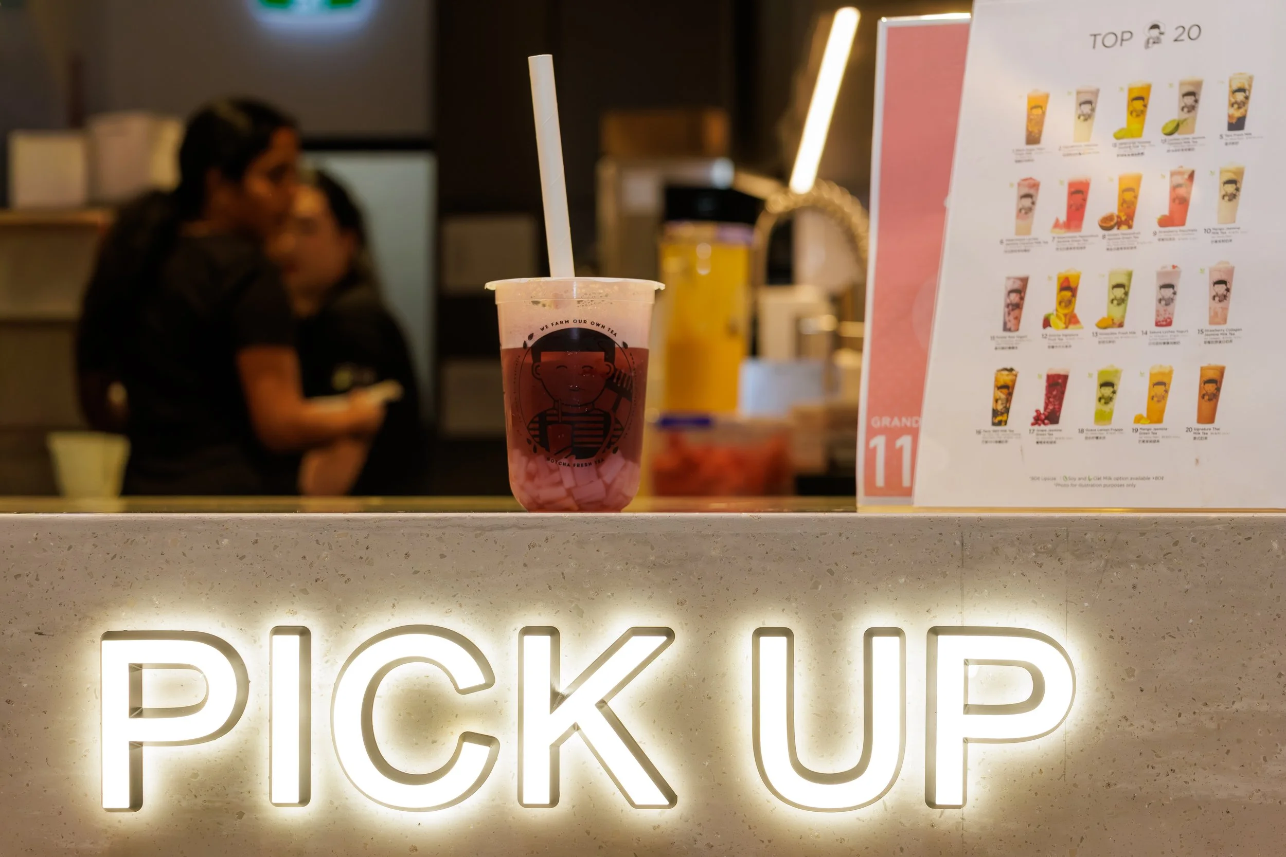 A pink bubble tea drink with a white straw on a counter, with a blurred menu and a person in the background.