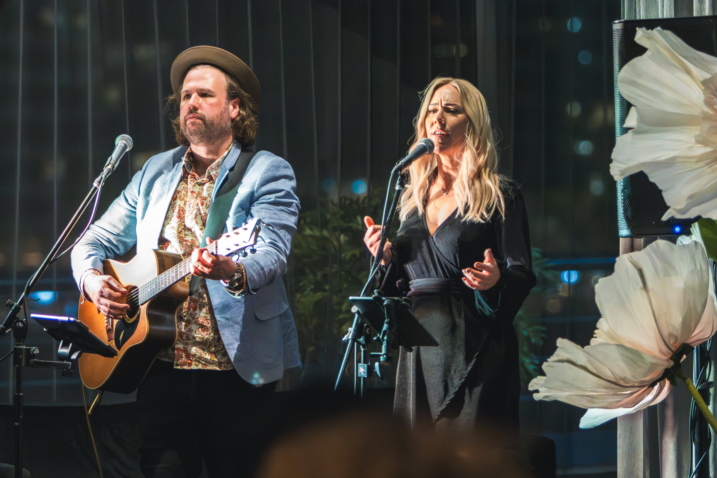 Two musicians perform on stage, one playing an acoustic guitar and the other singing into a microphone, with dark vertical background and flower decorations.