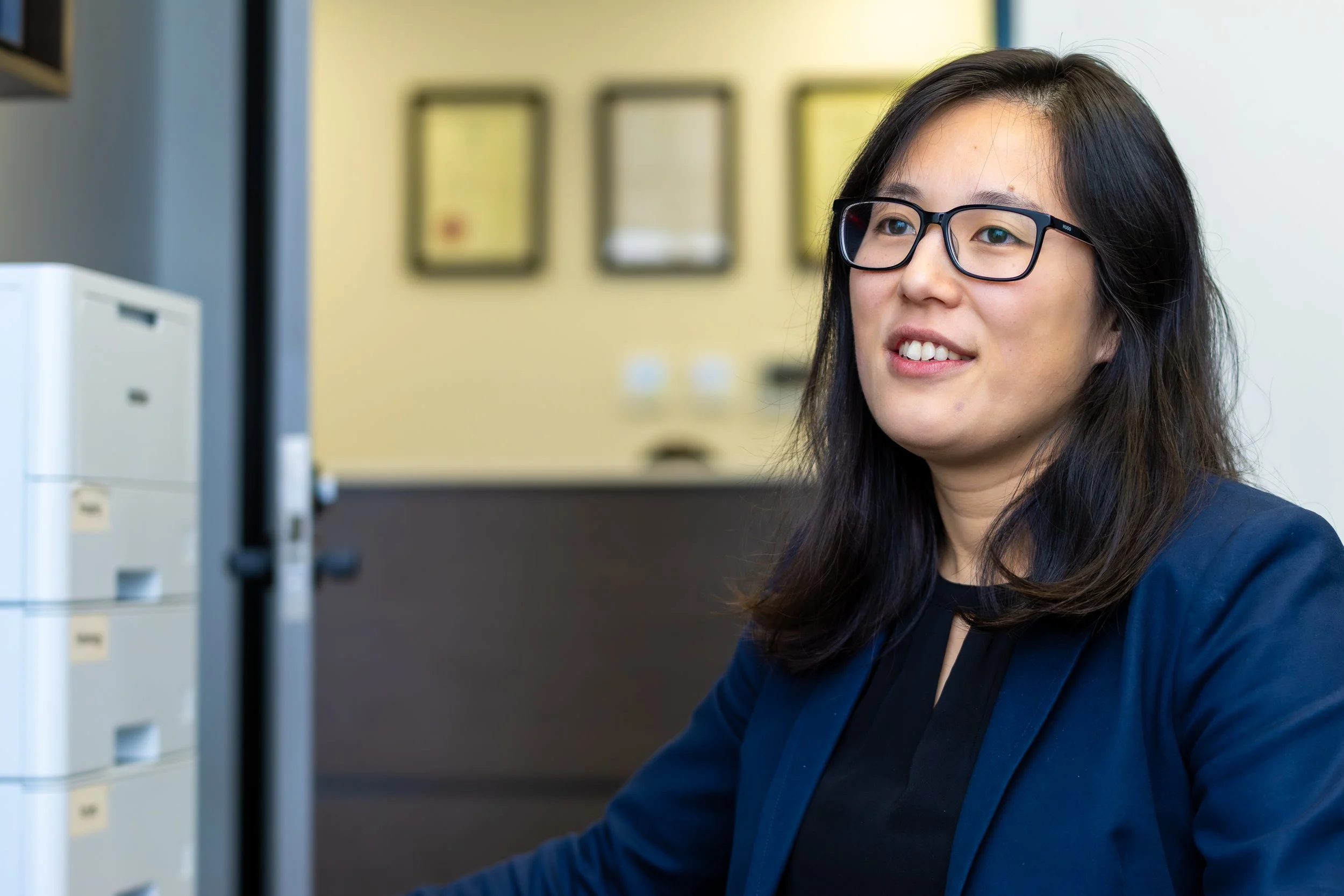A woman with black hair and glasses working at a desk in an office environment.