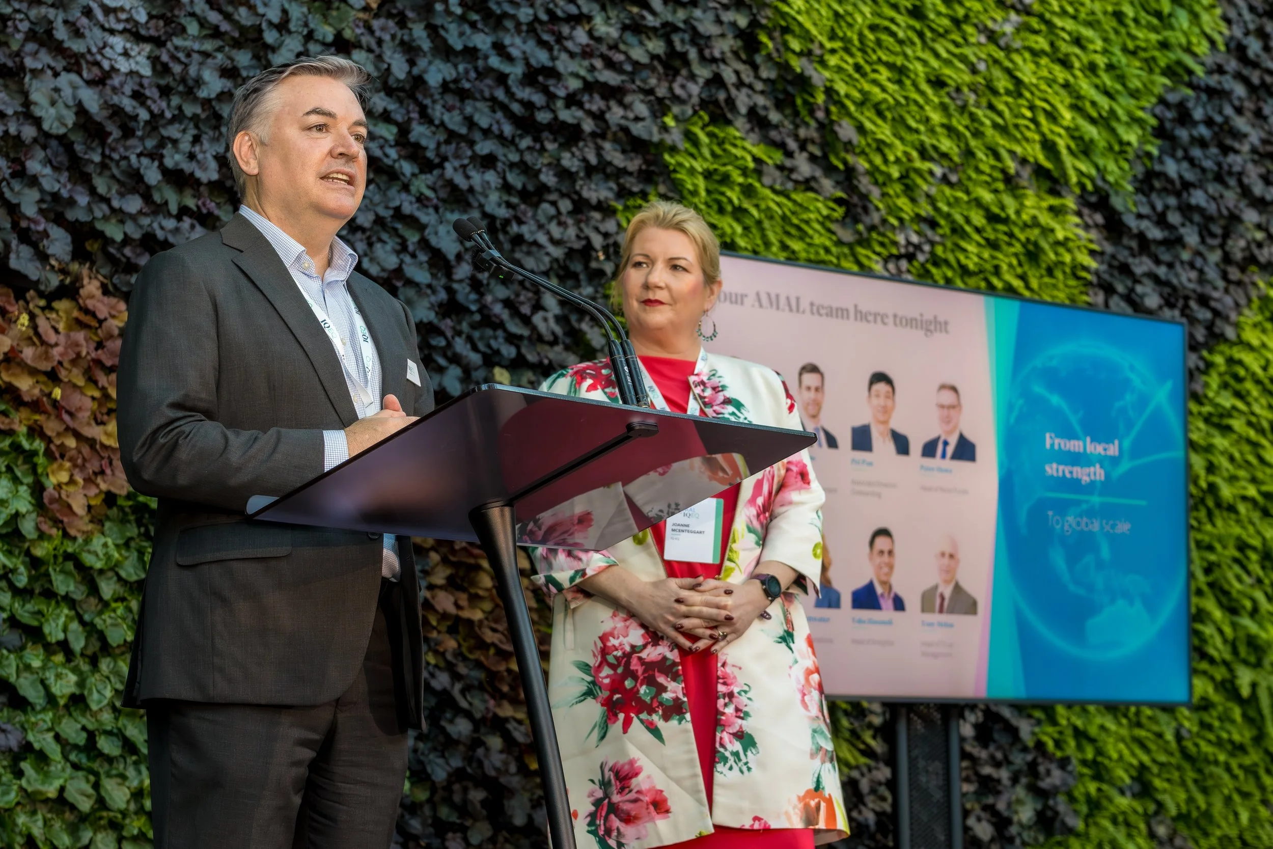 A man and a woman speaking at a podium during an outdoor event. The man is dressed in a dark suit and white shirt, while the woman wears a floral blazer over a red top. Behind them, a large screen displays text and images, and a wall of green foliage