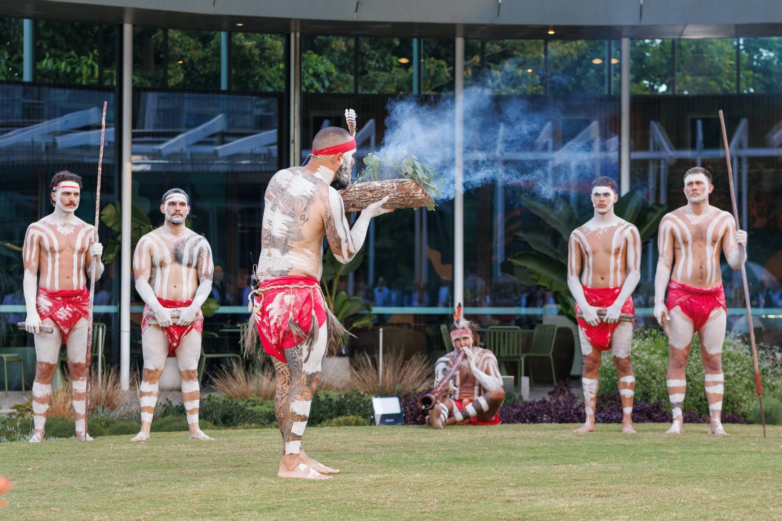 Indigenous performers dressed in traditional attire and body paint performing a cultural dance outdoors, with some holding spears and others sitting or lying on the grass.