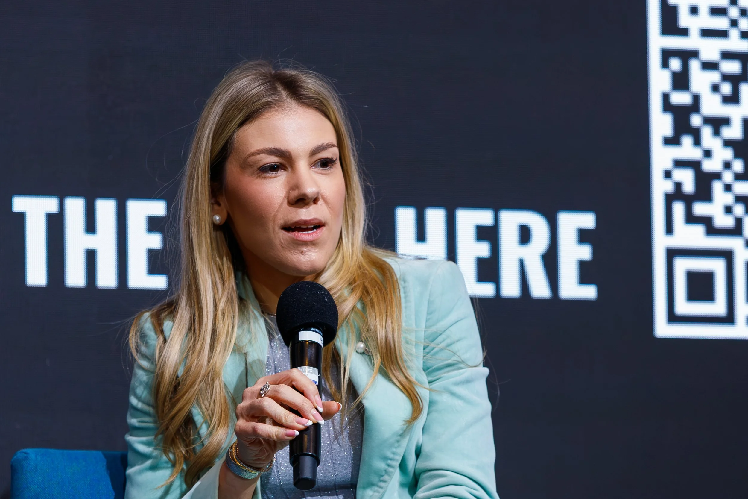 A woman with long blonde hair speaking into a microphone at an event, with a digital display behind her showing text and a QR code.