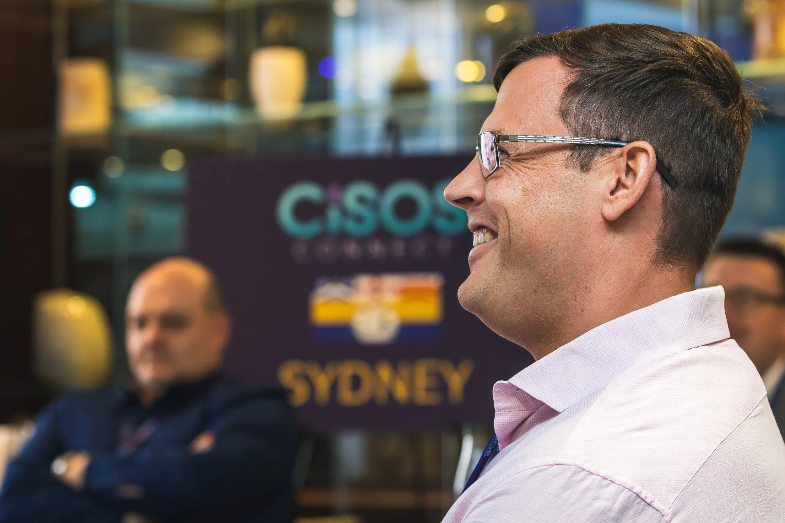 A man with short brown hair and glasses smiling, seen in profile, in a professional setting with blurred people and a sign reading 'CISO' in the background.