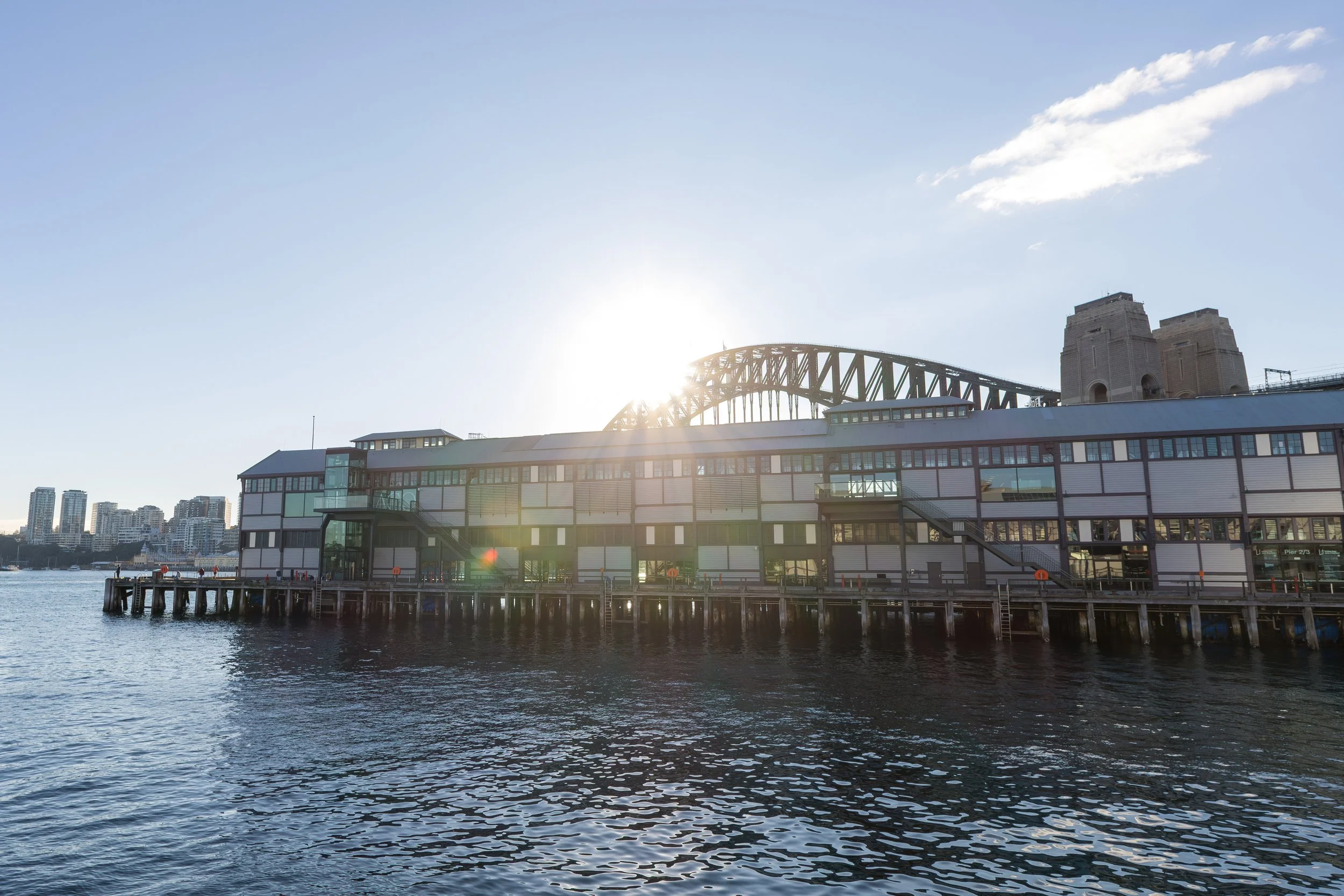 A waterfront view featuring a modern building on a pier with the Sydney Harbour Bridge in the background, sunlight shining through, and a clear blue sky.