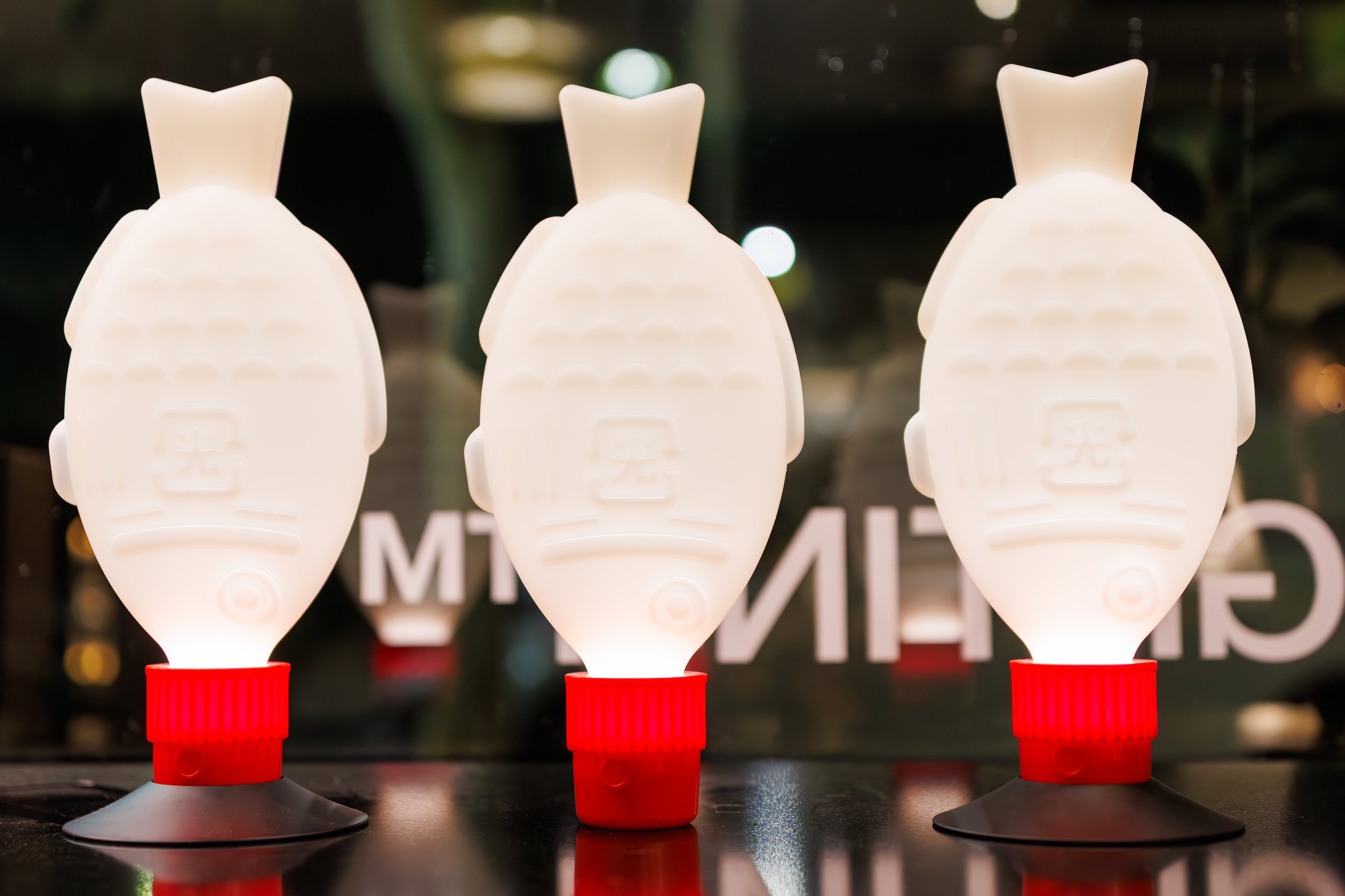 Three white glue bottles with red caps arranged in a row on a black surface, with a dark background containing blurred lights.