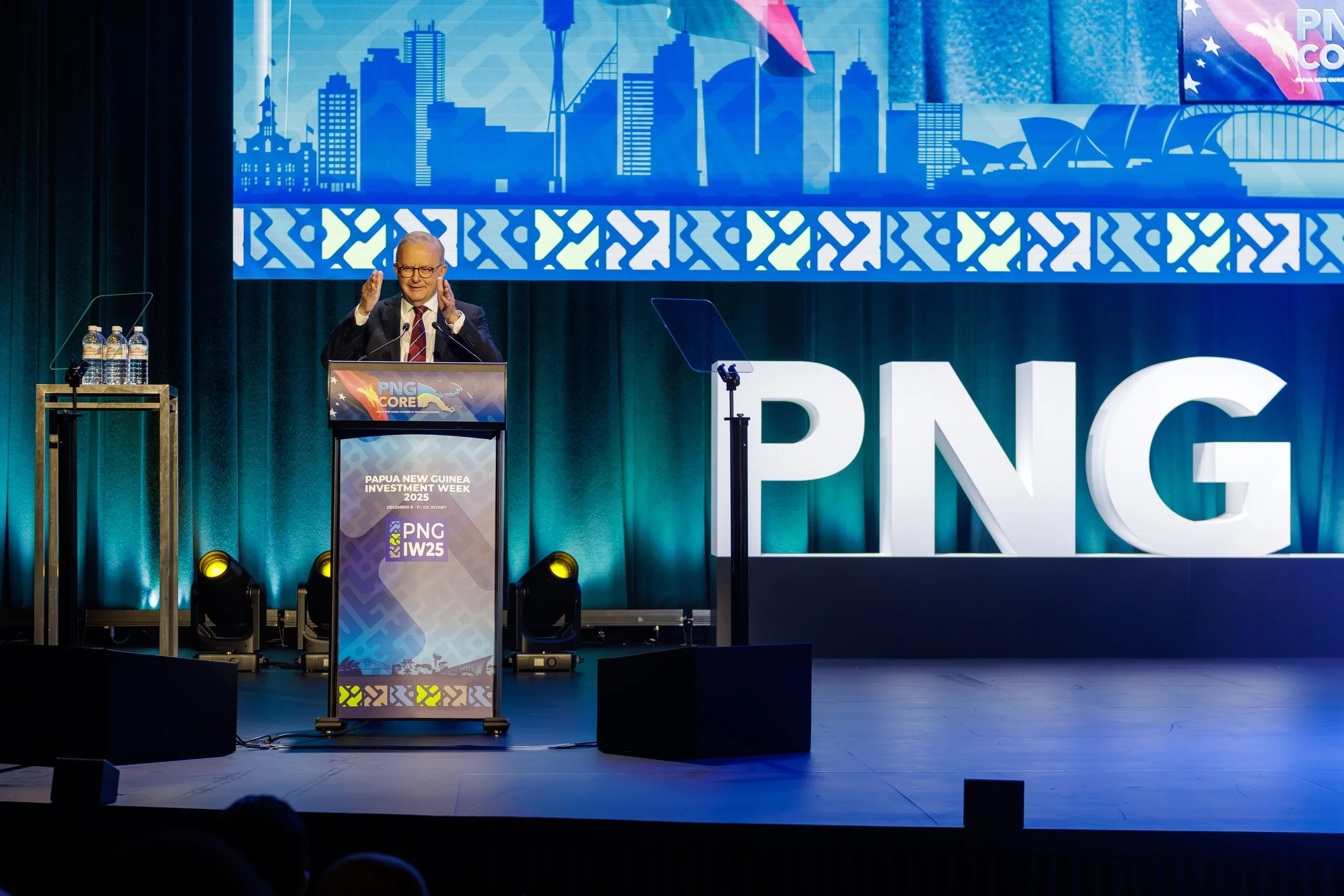 A man in a suit speaking at a podium during Papua New Guinea Investment Week 2025 conference. Behind him is a large screen displaying a blue map of a city skyline with modern buildings, the PNG logo, and the event title. There are water bottles on a 