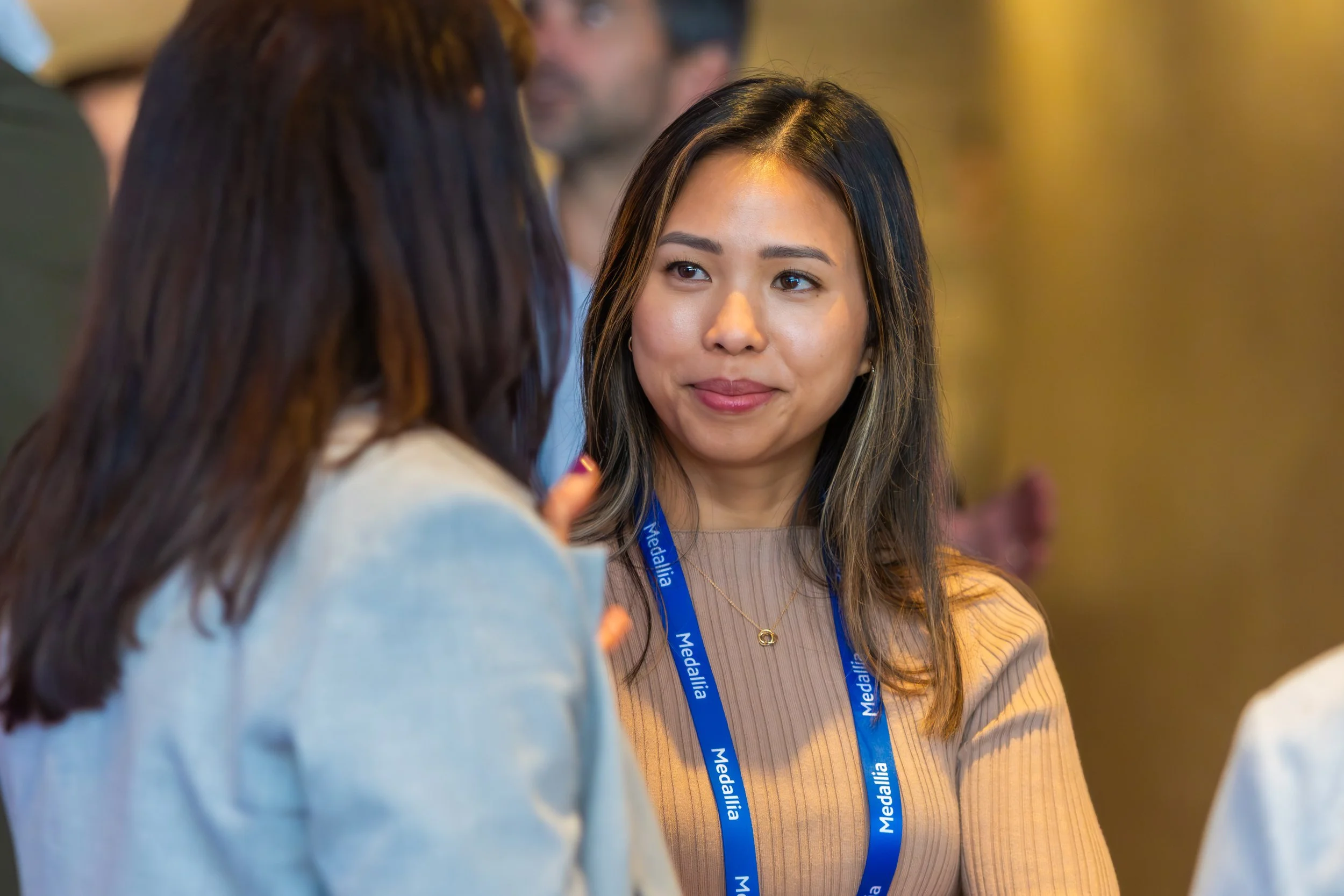 A woman with long dark hair and a beige top smiling while talking to a person with long dark hair wearing a white top. The woman is wearing a blue lanyard.