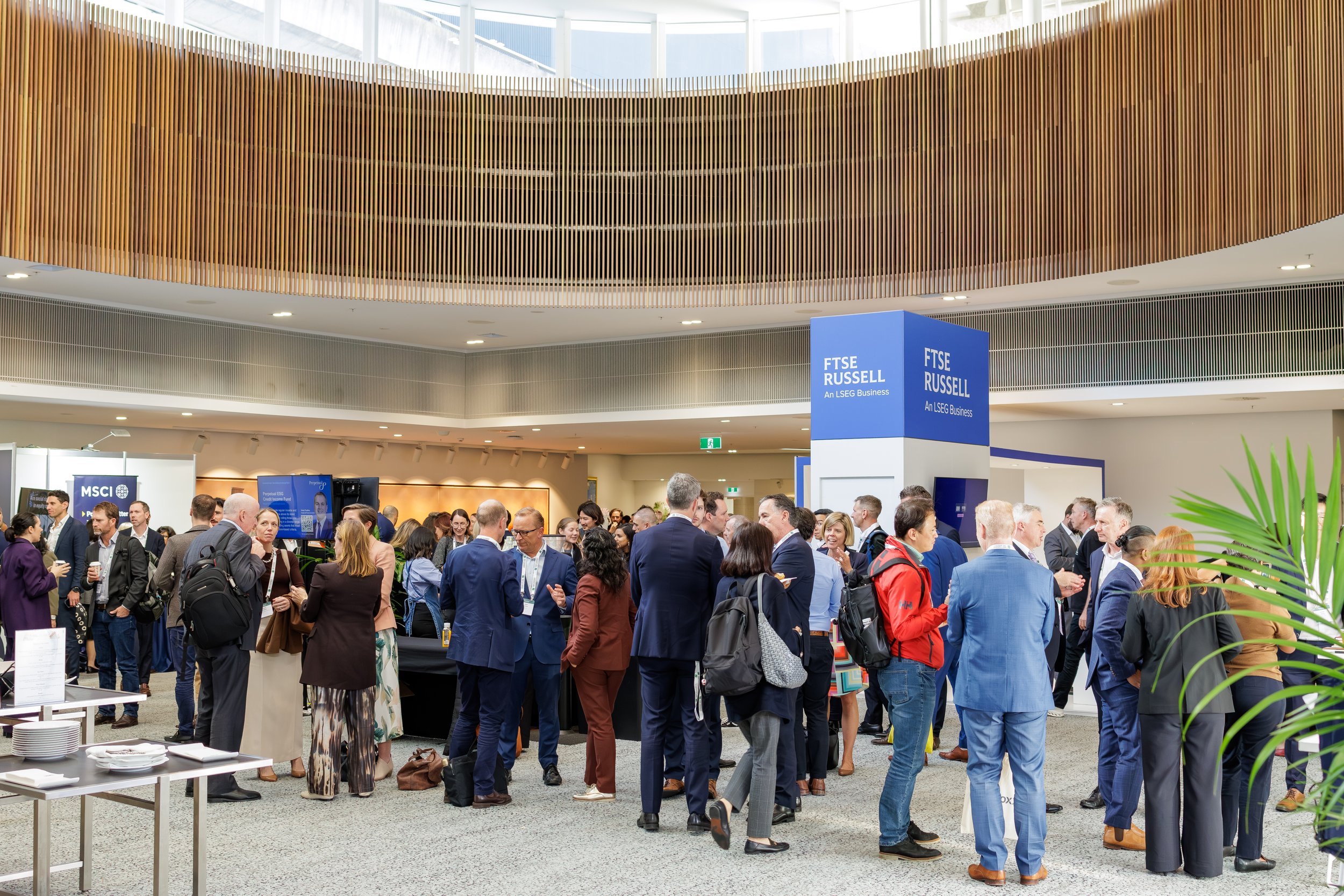 A conference or trade show with many attendees gathered in a large, modern indoor space. There are display booths, including one labeled 'FTSE RUSSELL,' and people dressed in business attire are engaged in conversations. The building has a high ceili