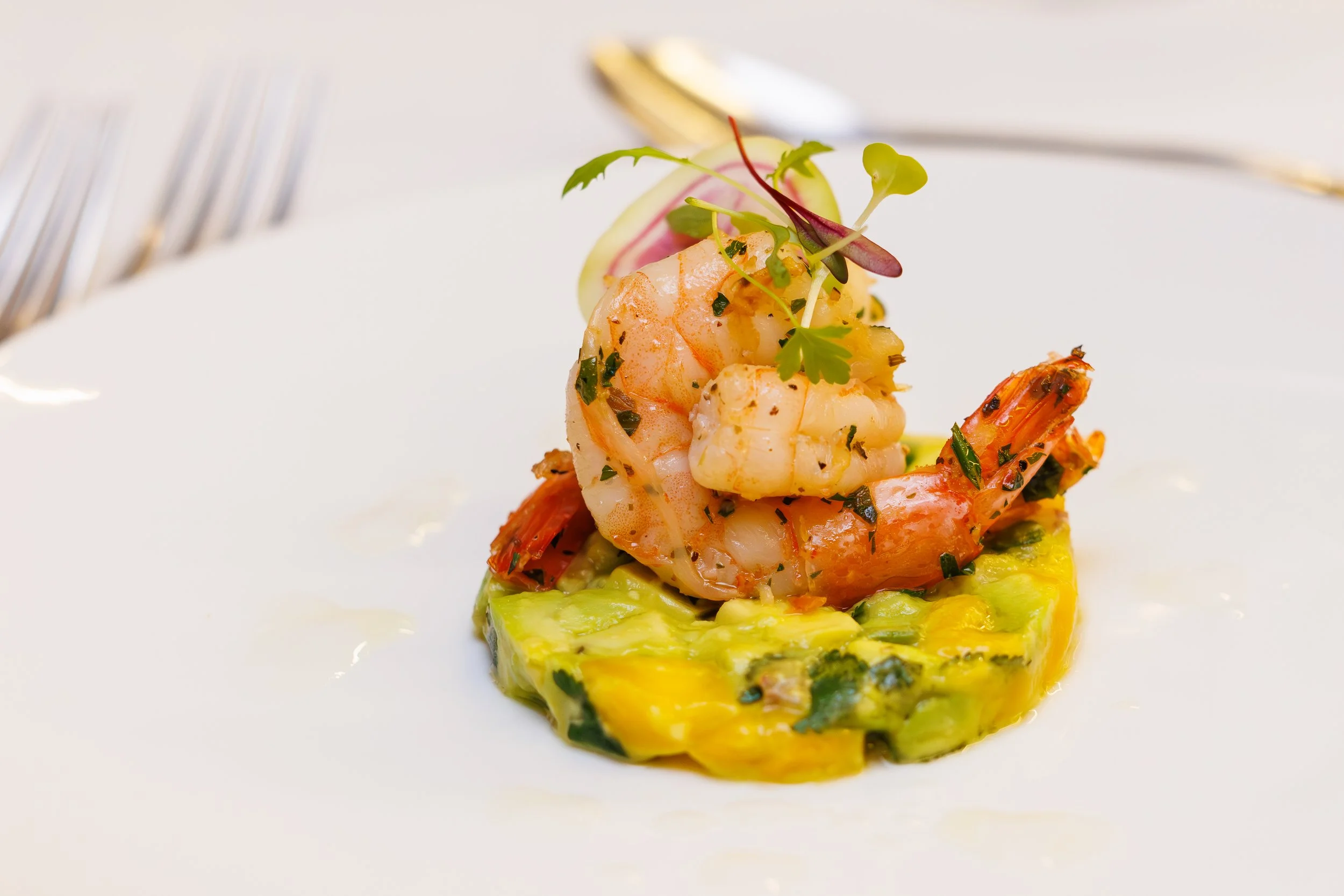 Close-up of a gourmet dish featuring shrimp on a bed of green avocado or zucchini mixture, garnished with microgreens, on a white plate with a fork and knife in the background.