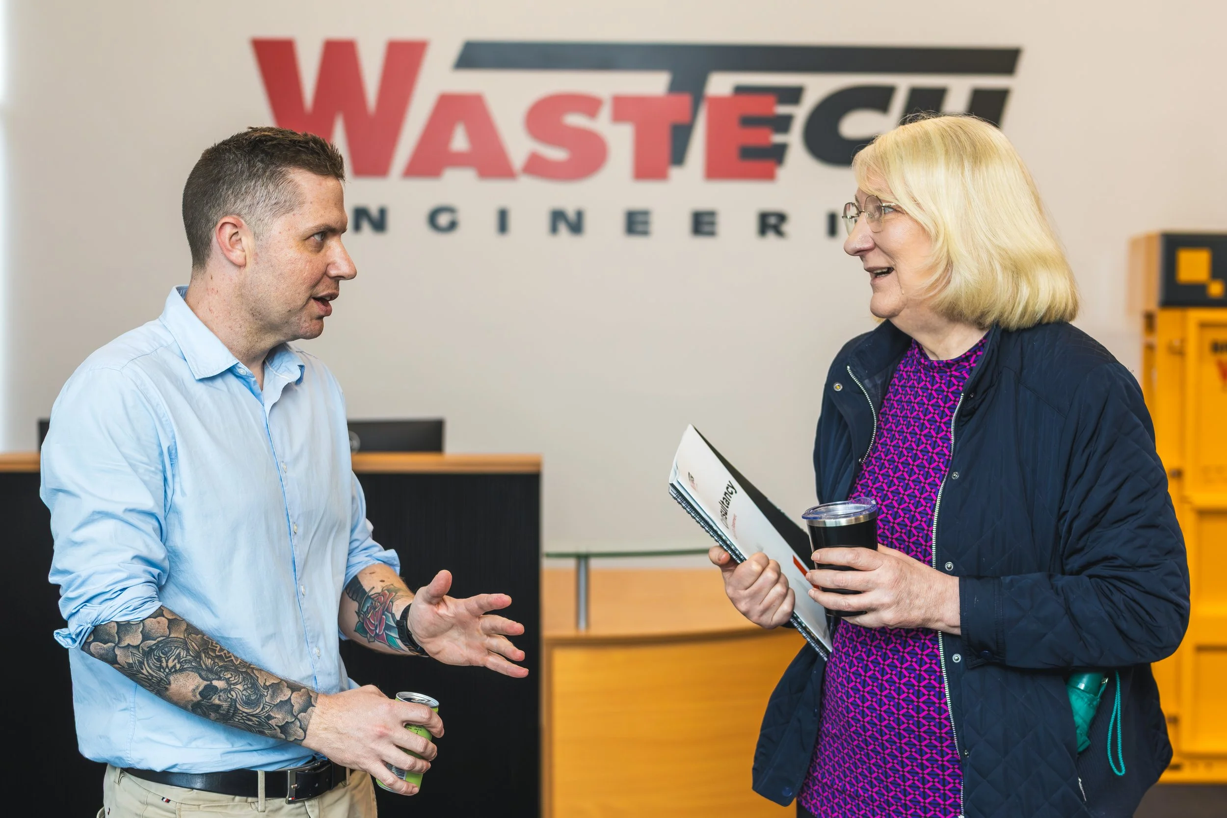 Two individuals engaged in conversation inside an office or industrial space, with a large 'WASTE' sign in the background. The man on the left, with tattoos on his arms and a light blue shirt, is speaking and gesturing with his hand. The woman on the