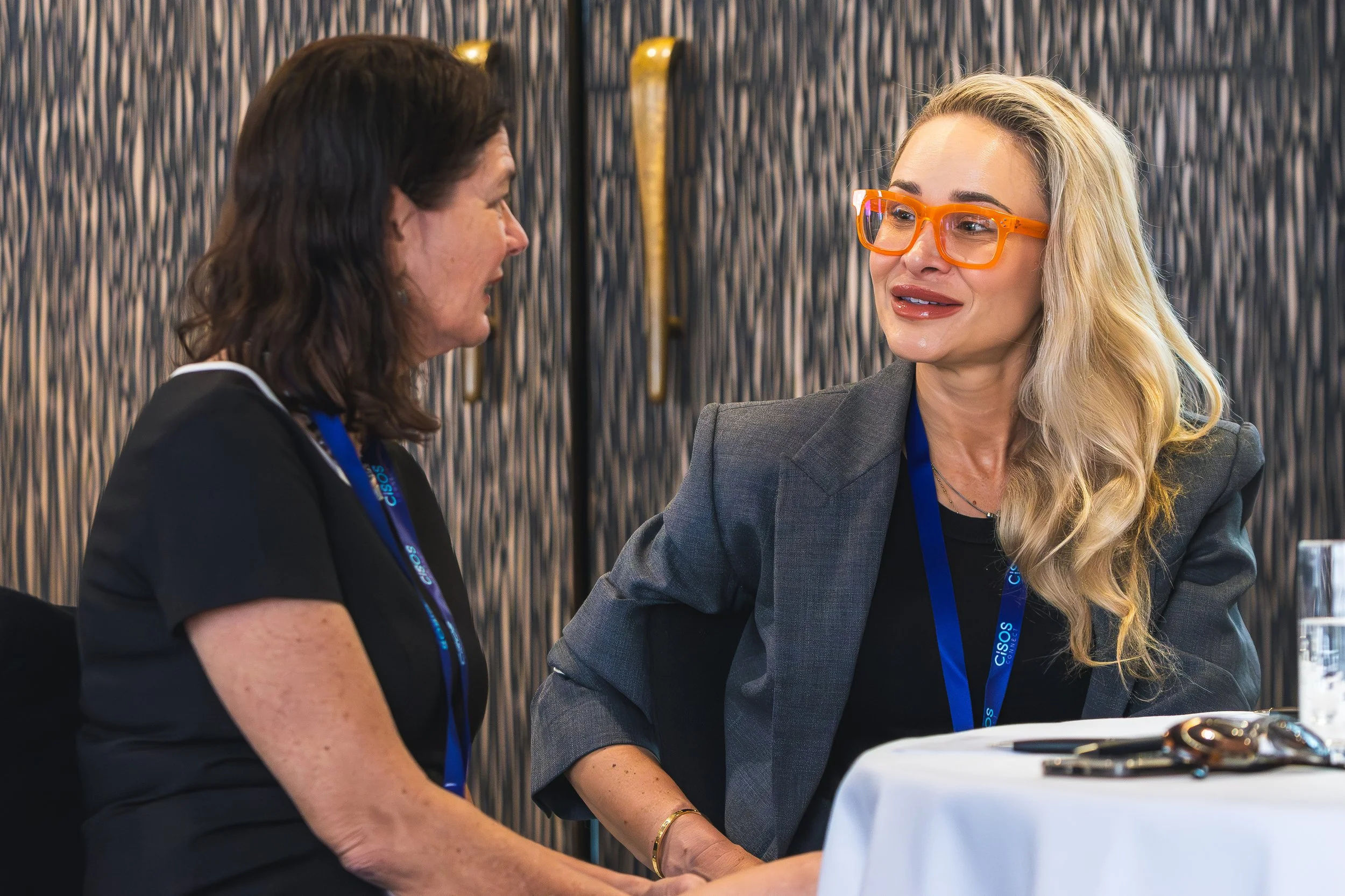 Two women talking at a conference, one wearing orange glasses and gray blazer, the other with dark hair in a black blazer, seated at a table with conference badges around their necks.