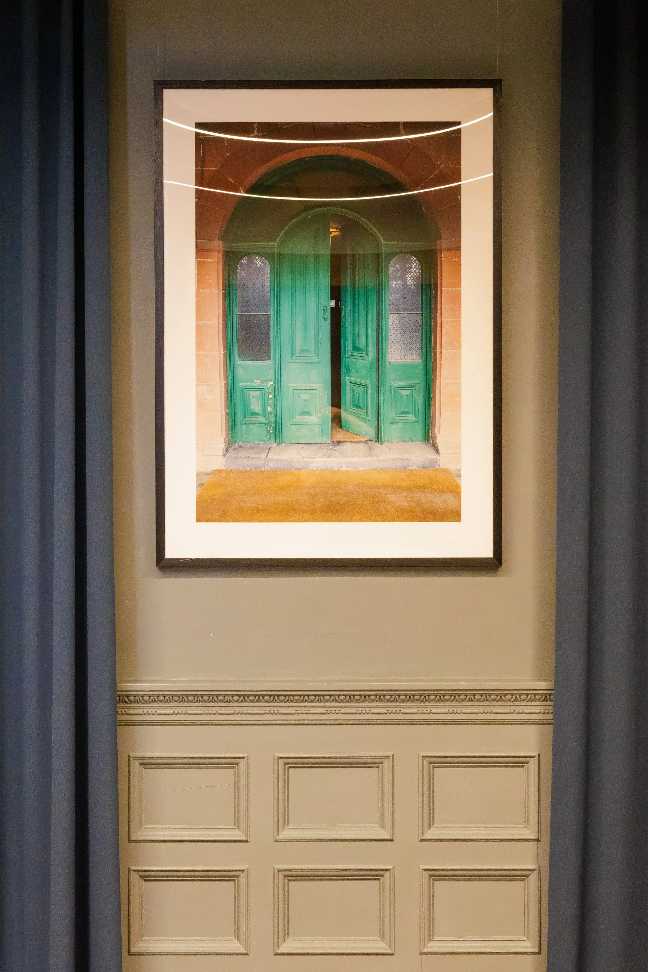 A framed photograph of a green door with arched top windows, placed on a white wall with ornate molding and flanked by dark curtains.