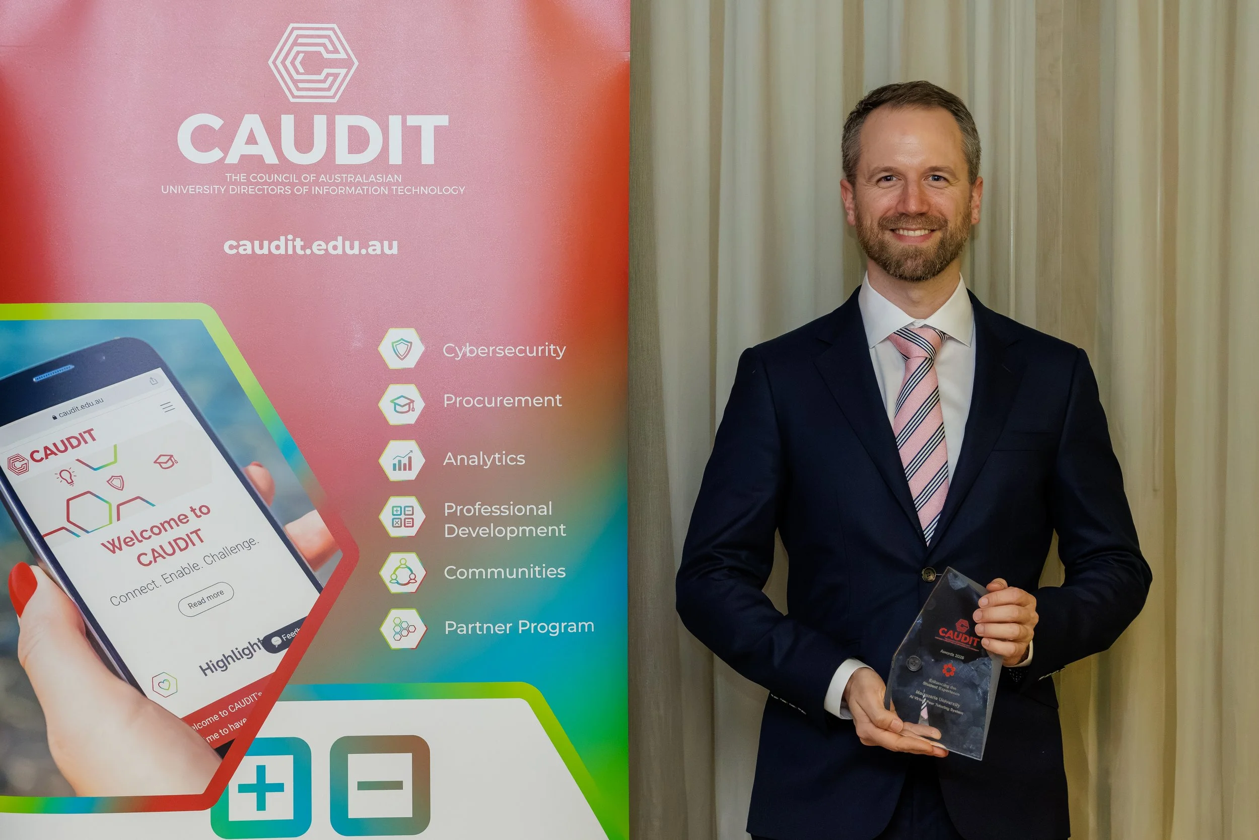 A man in a dark suit and pink striped tie standing beside a CAUDIT banner, holding an award, smiling at the camera.